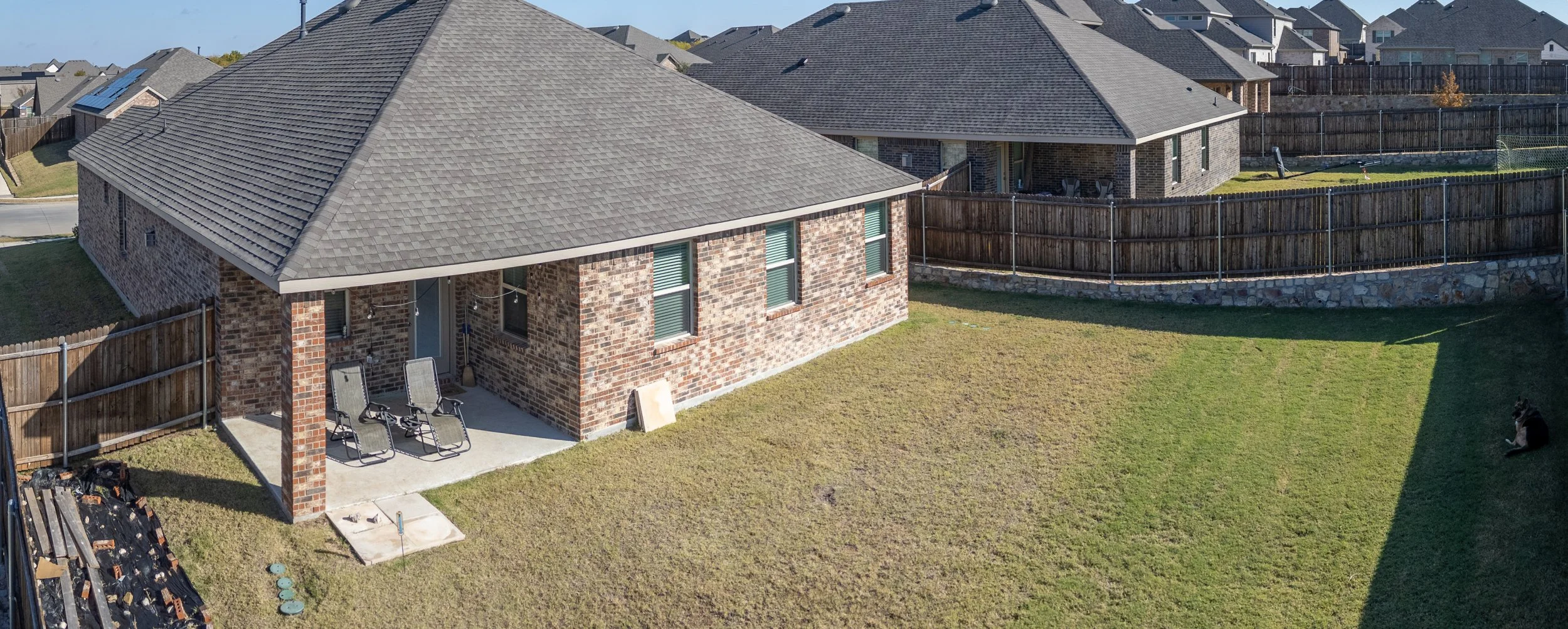 Backyard of a brick house with lawn, patio furniture, and a dog lying on the grass, fenced yard, neighboring houses in the background.