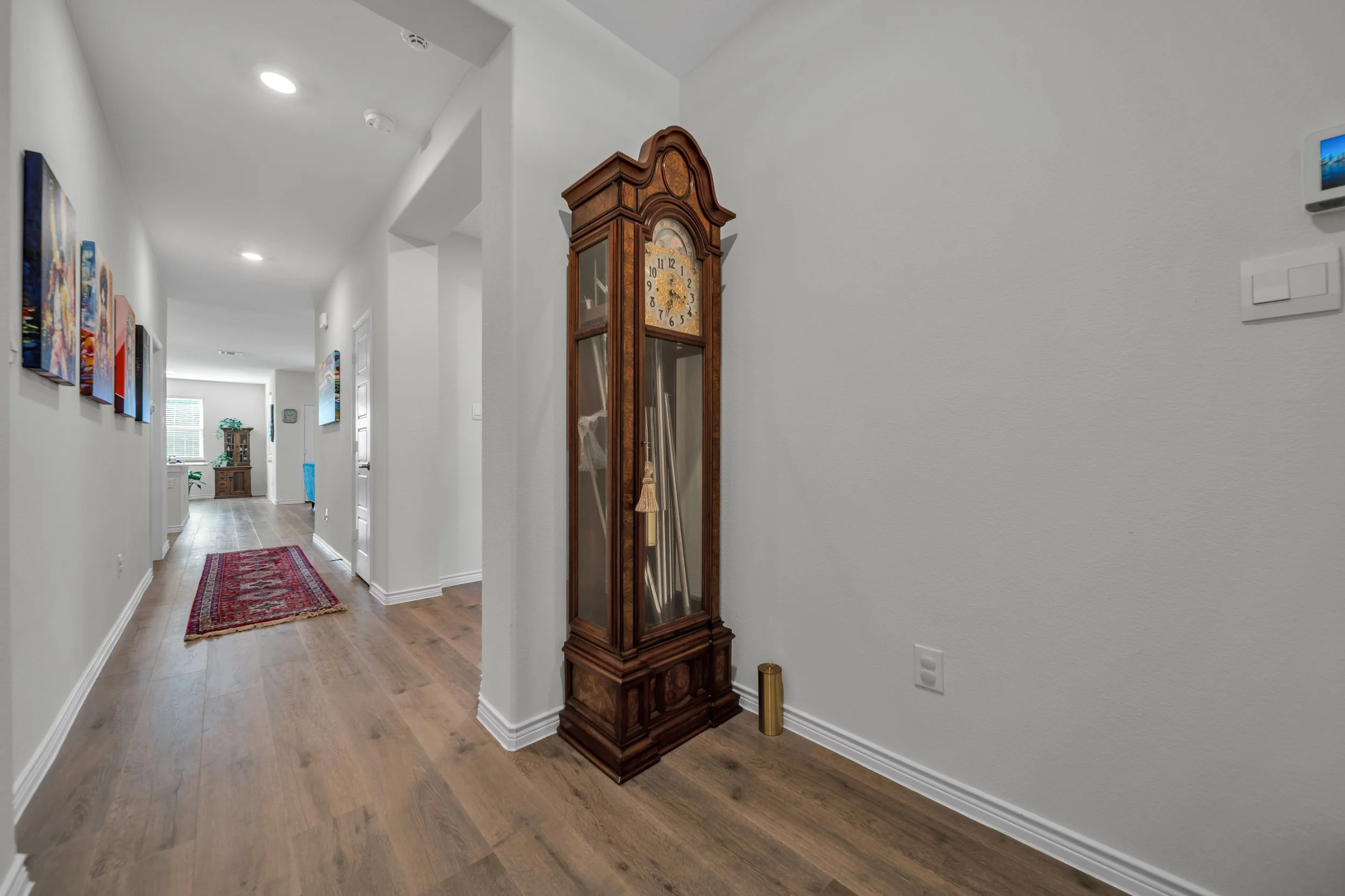 A hallway with wooden flooring, colorful paintings on the wall, an antique grandfather clock, and a red patterned rug.