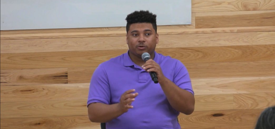Justin Parker in a purple shirt speaking into a microphone against a wooden background.