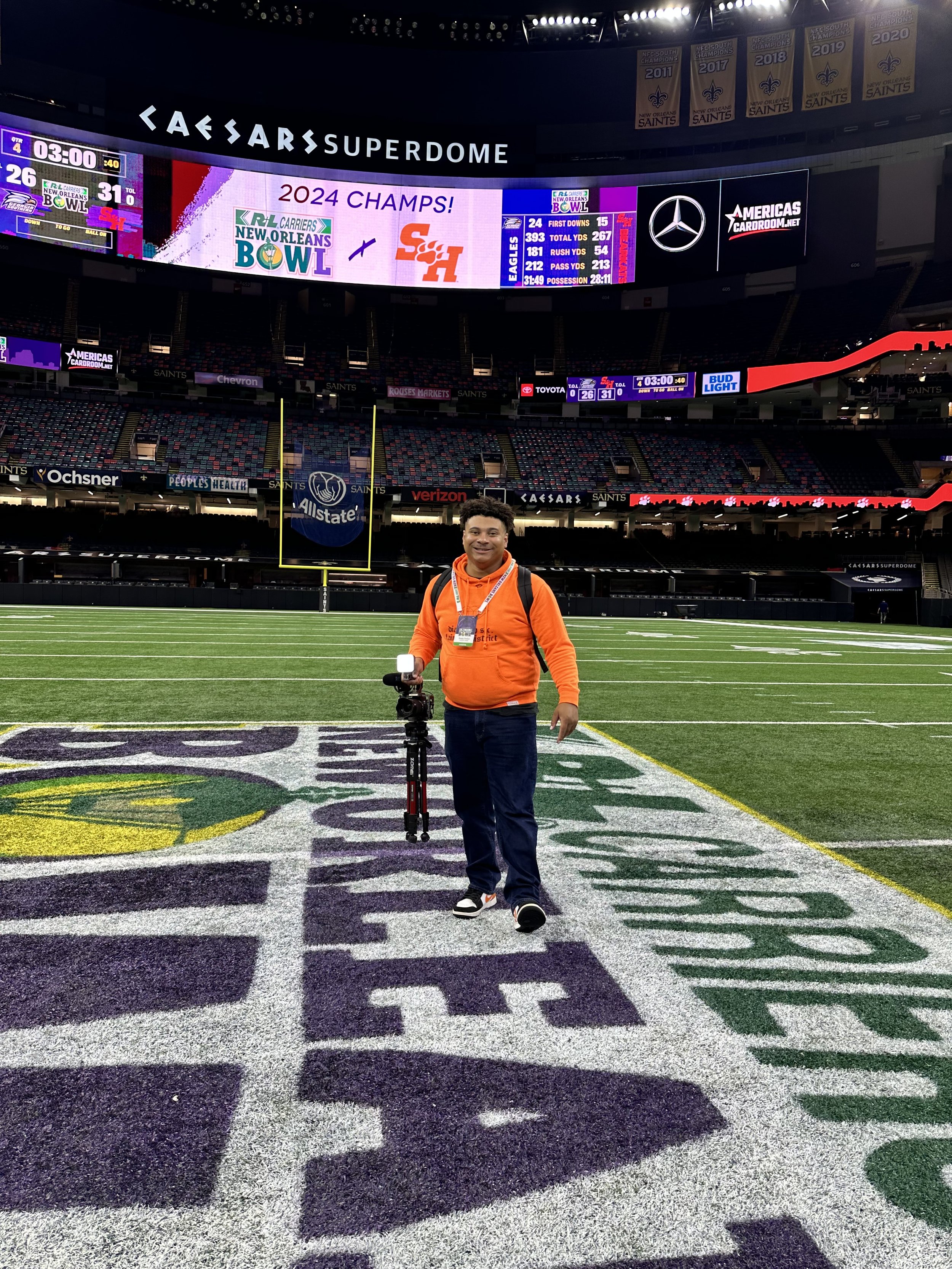 Justin Parker in an orange hoodie stands on a football field at the Caesars Superdome. The scoreboard displays "2024 CHAMPS!" and team scores. The field is marked for the New Orleans Bowl. He is holding a camera on a tripod and smiling at the camera.