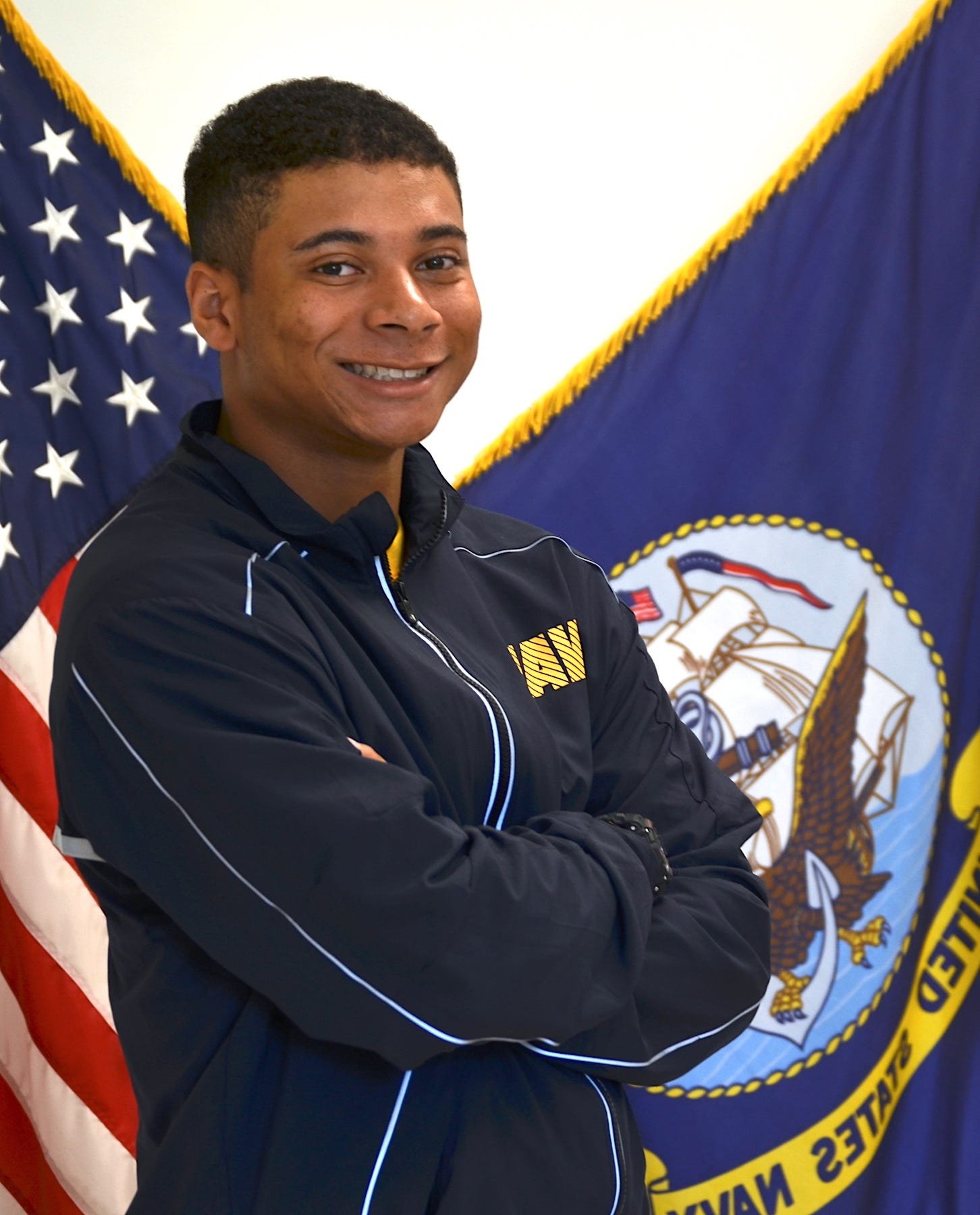 Justin Parker in Navy attire standing with arms crossed, smiling in front of American and Navy flags indoors.