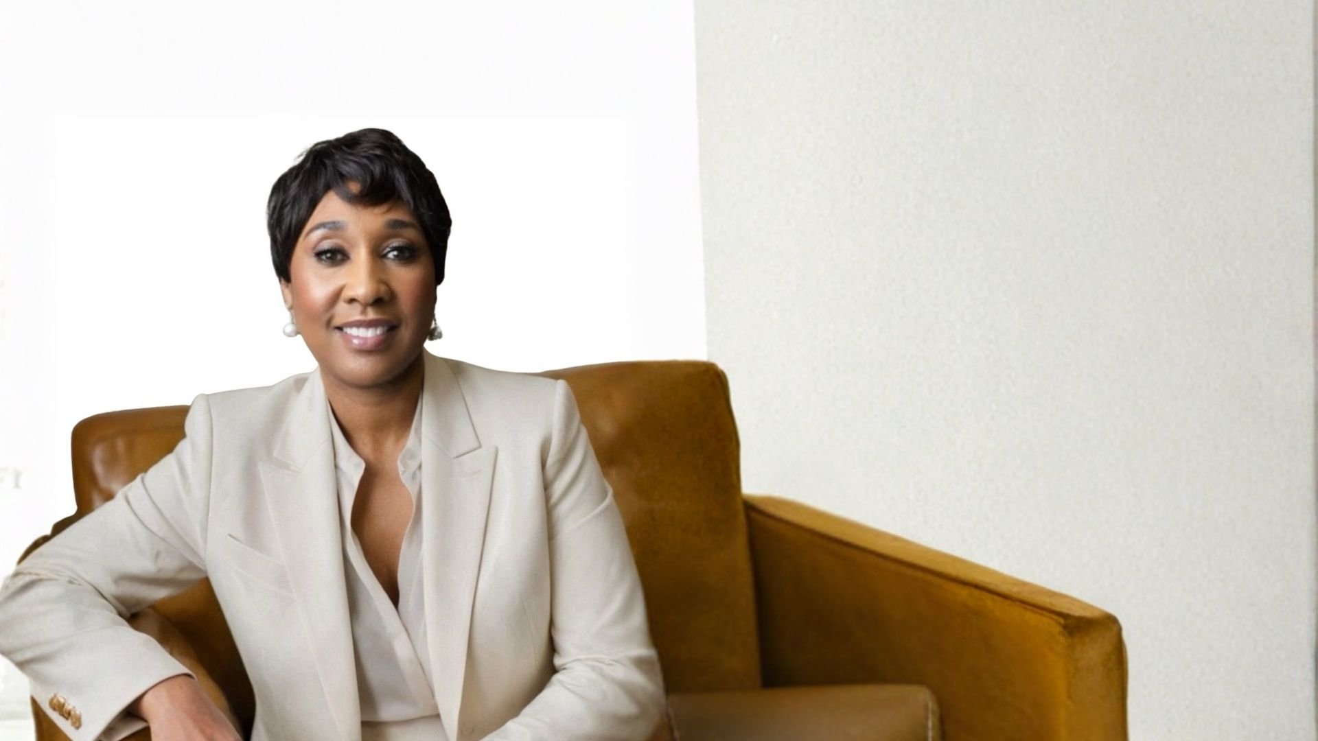 A woman with short black hair, wearing a cream-colored blazer, sitting on a brown leather couch in a minimalistic room with white walls, smiling at the camera.