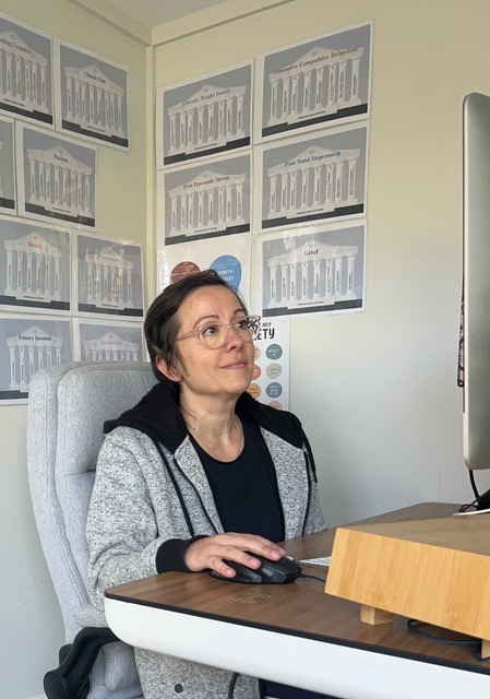 A woman with short dark hair and glasses sitting at a desk, using a computer mouse in an office with charts on the wall behind her.