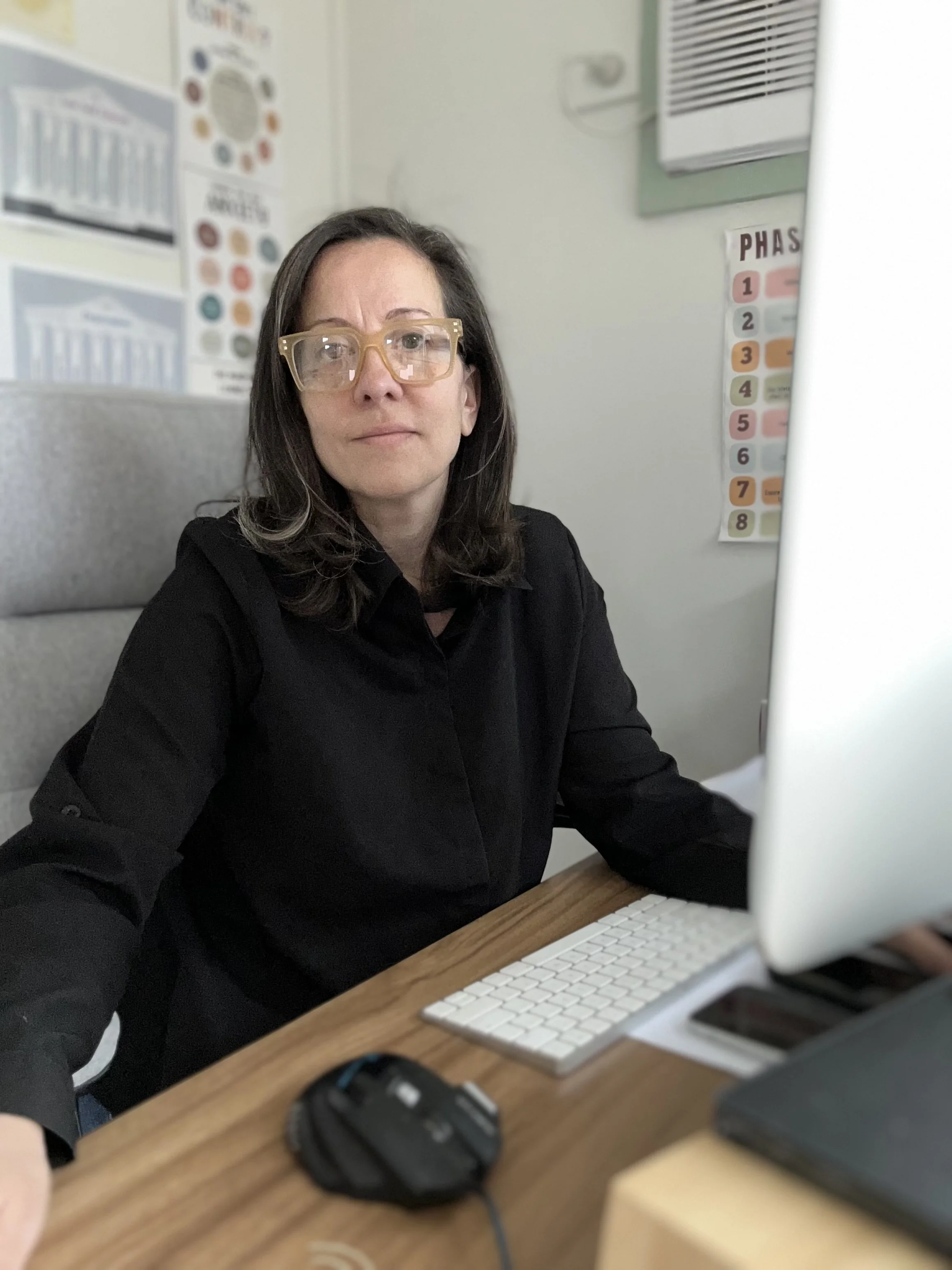 A woman with shoulder-length dark hair, wearing beige glasses and a black shirt, sitting at a office desk with a computer monitor, keyboard, mouse, and phone.