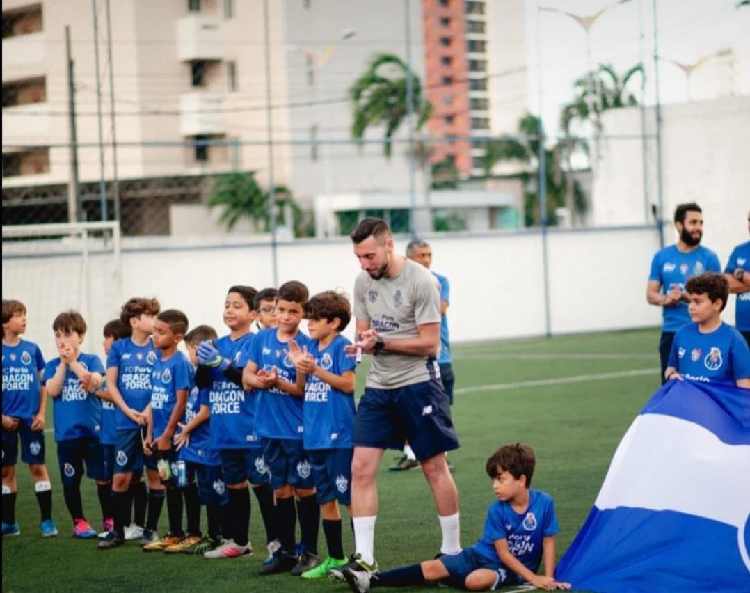 A group of young boys in blue sports uniforms with 'FC Porto Dragon Force' on them, standing on a soccer field. A coach or adult is talking to them, with a few other adults nearby. The setting appears to be an outdoor sports facility with tall buildings and trees in the background.