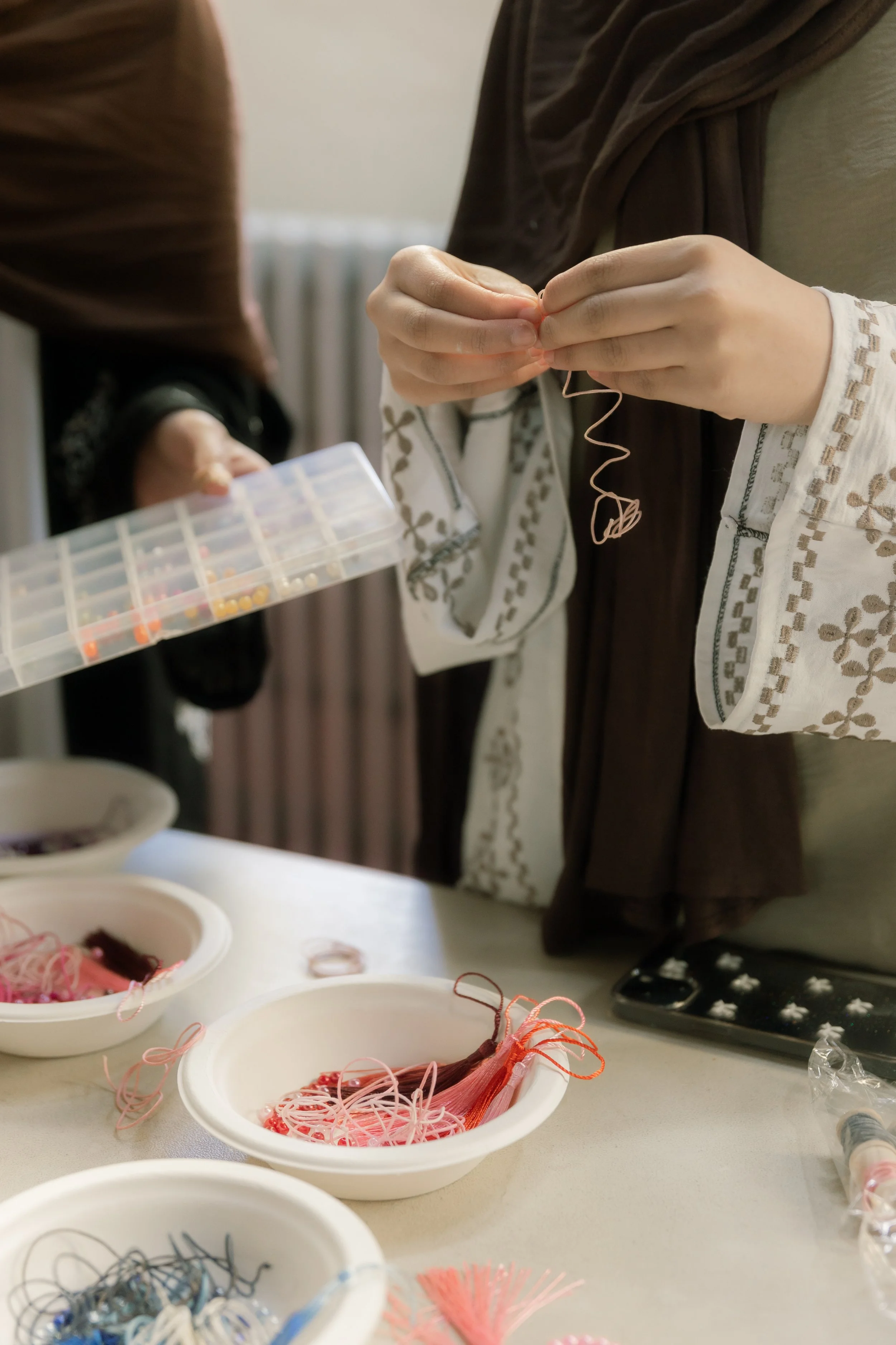 Close-up of hands crafting with strings and beads, bowls of colorful threads on a table.