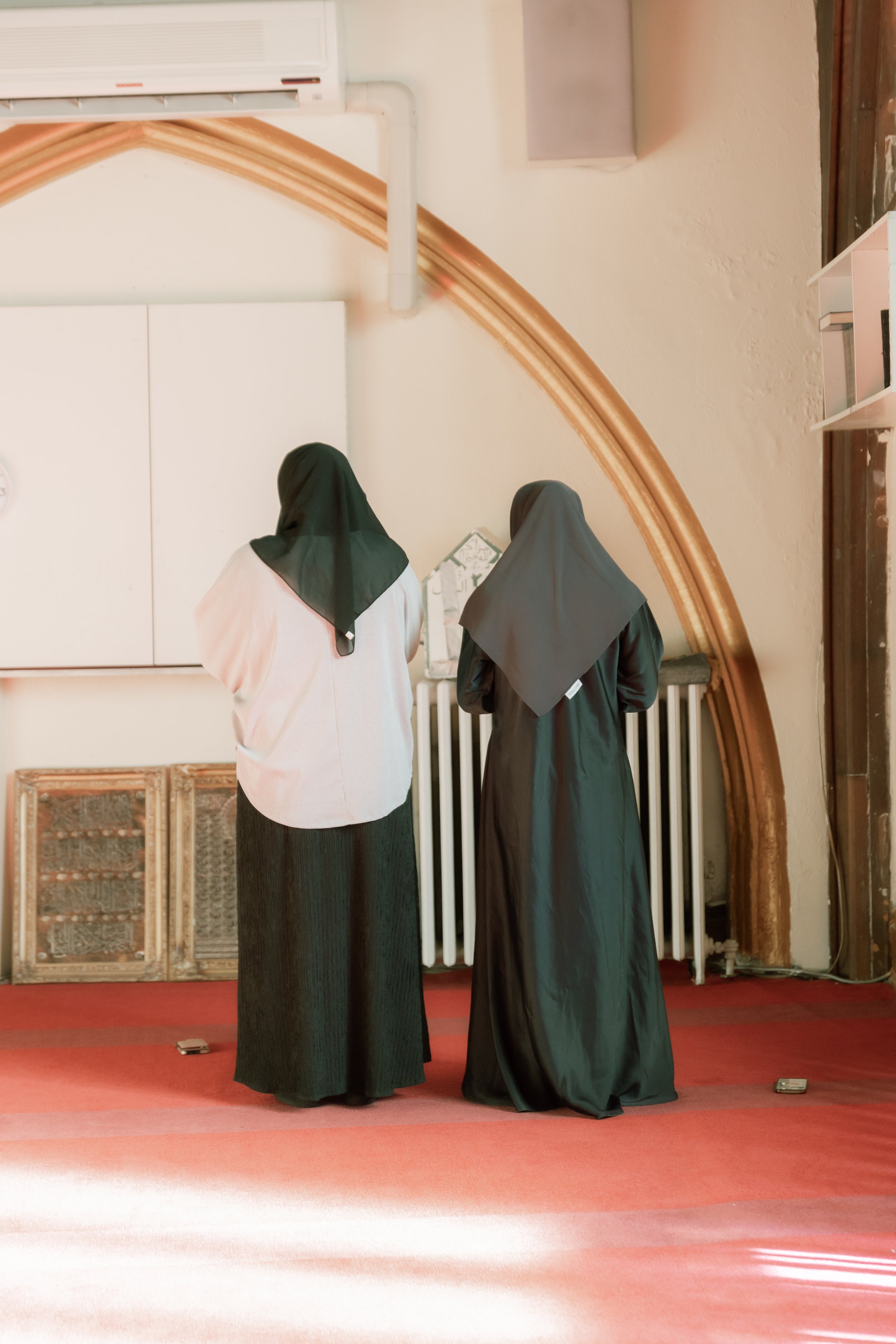 Two people wearing traditional garments standing on a red carpet in a room with arches and framed decorations.