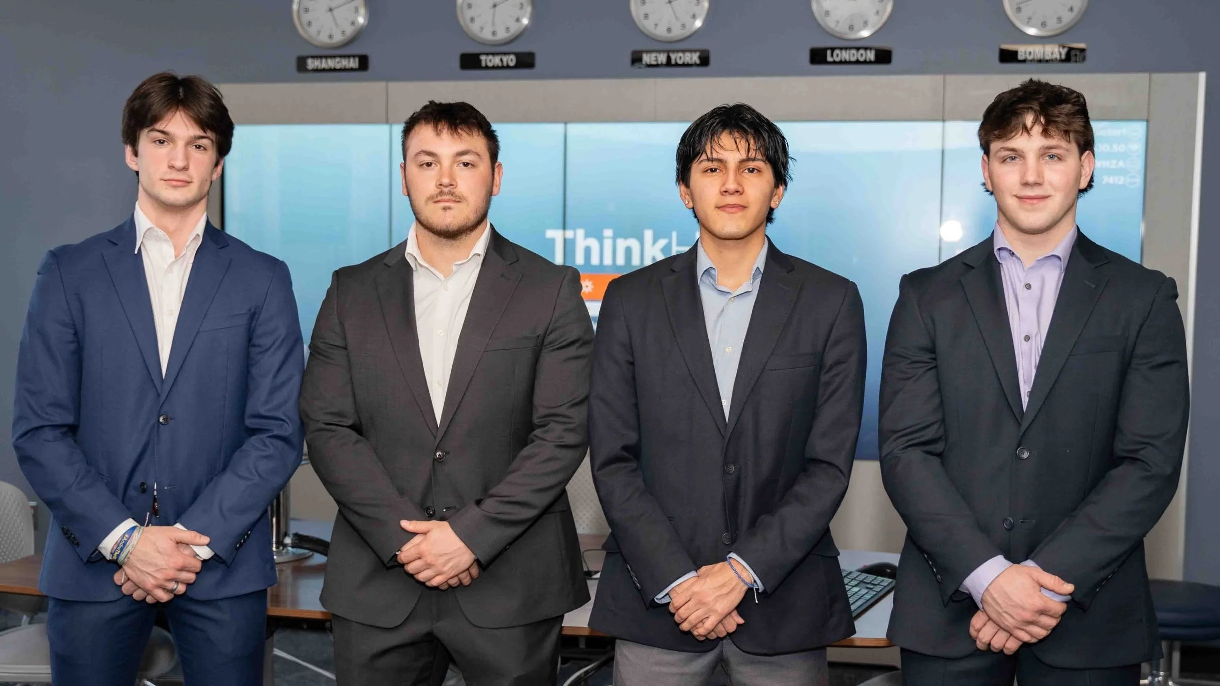Four young men in suits standing together in an office setting with clocks showing different time zones above them.