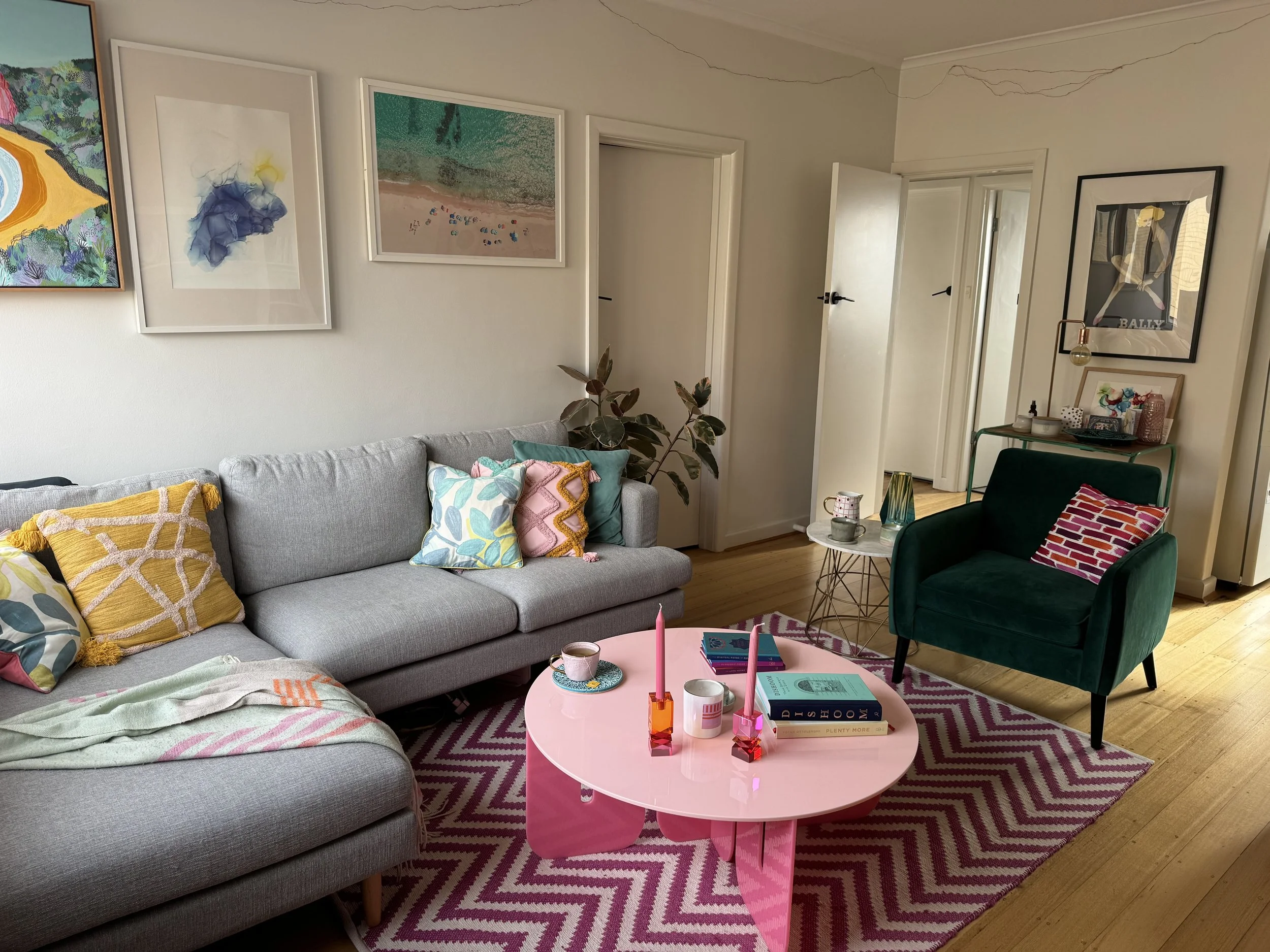 Cozy living room with a light gray sofa adorned with colorful pillows, a pink coffee table with candles and books, a dark green armchair with a striped pillow, a patterned rug, and wall art, with a side table and a plant.