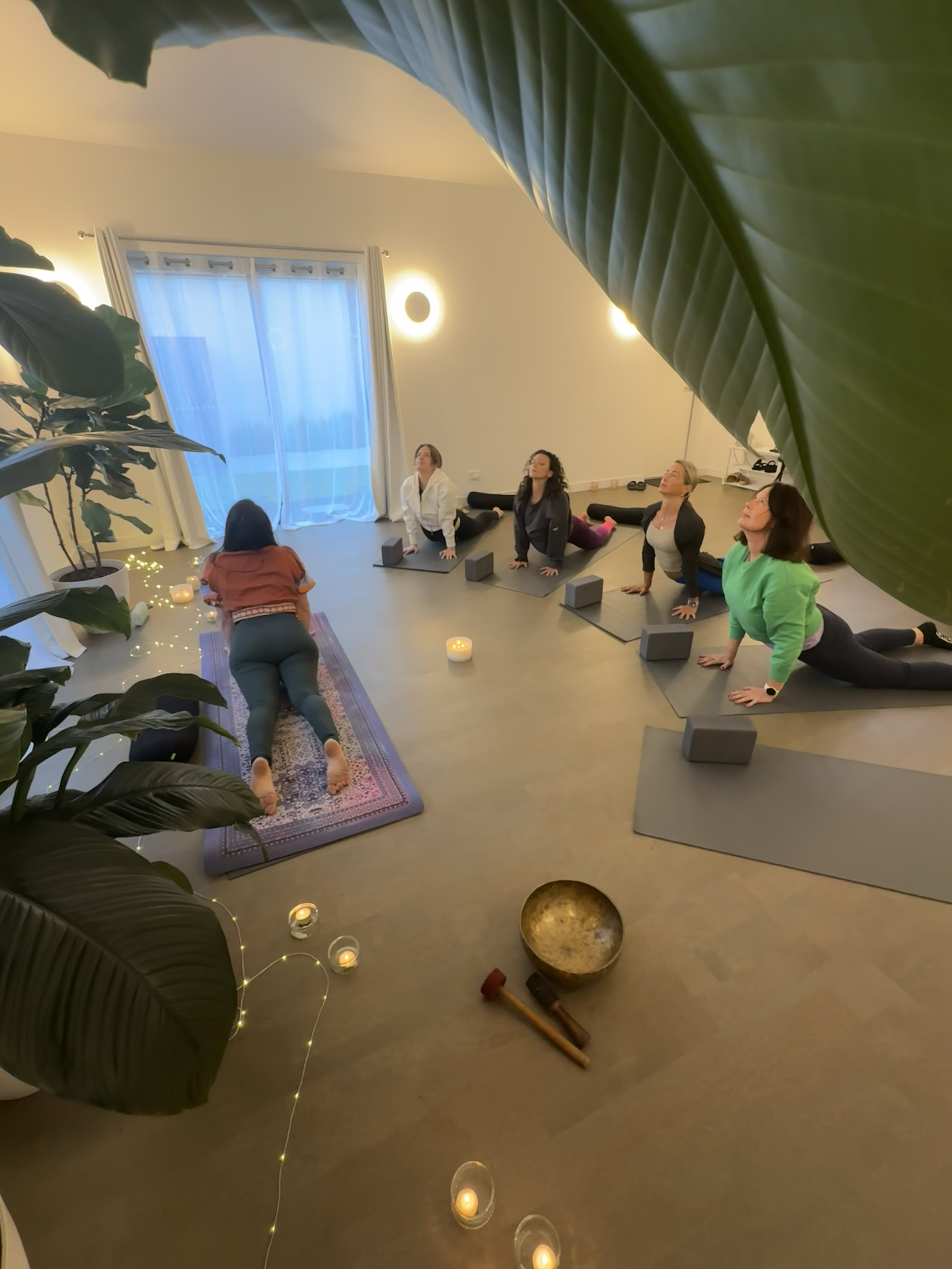 Group of people participating in a yoga class in a cozy, well-lit room with candles, plants, and a large window.