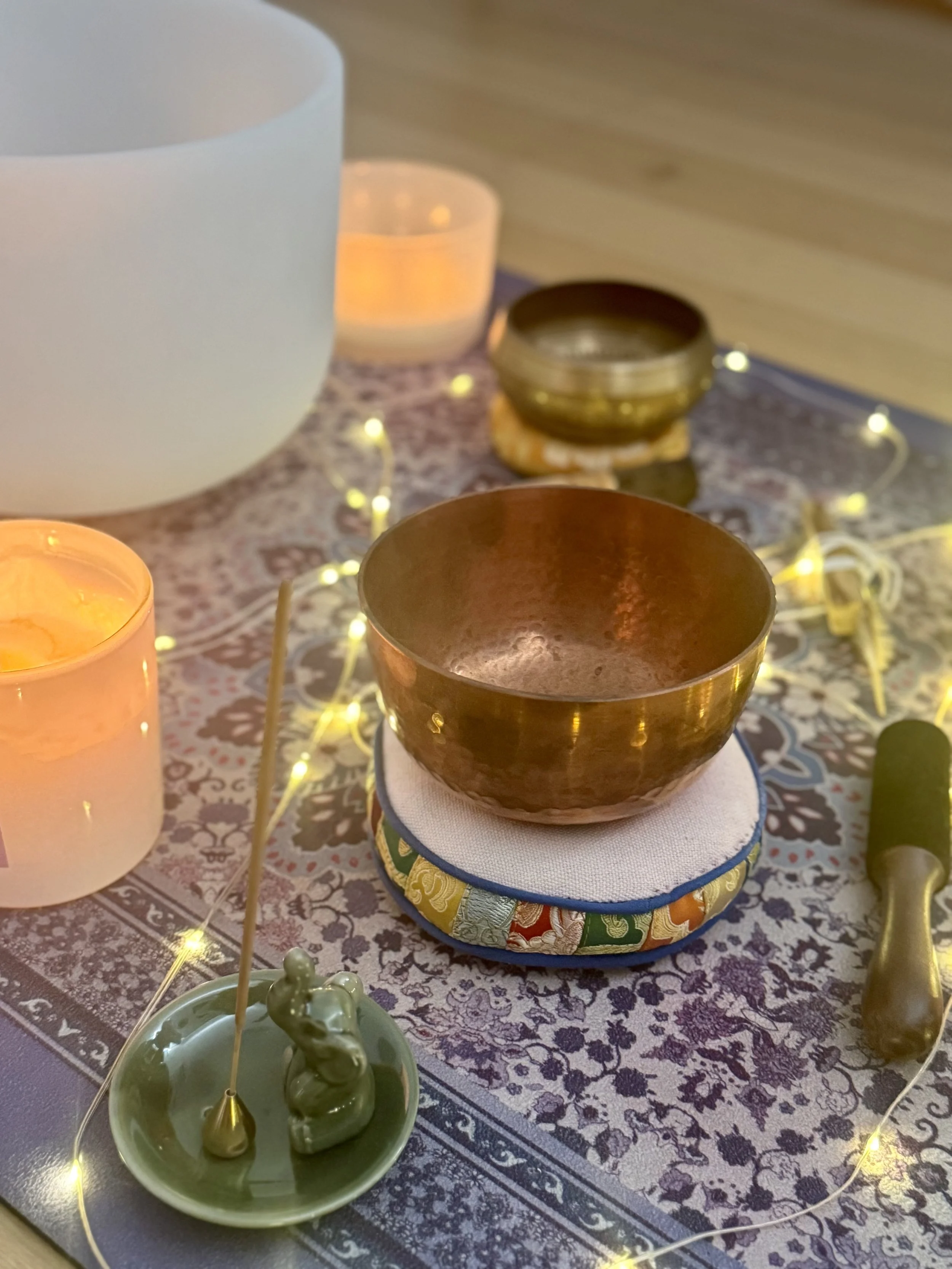 A meditation or spiritual altar setup with candles, a singing bowl, a small statue, and decorative items on a patterned cloth.