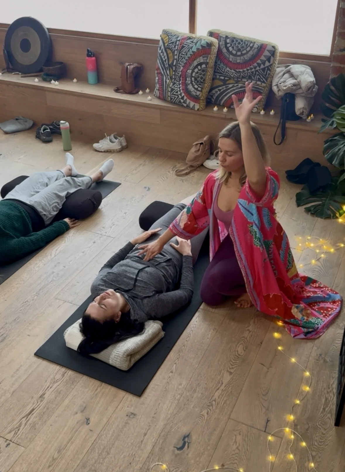A woman practicing Reiki on a person lying on a yoga mat in a cozy room with candles, pillows, and decorative blankets.