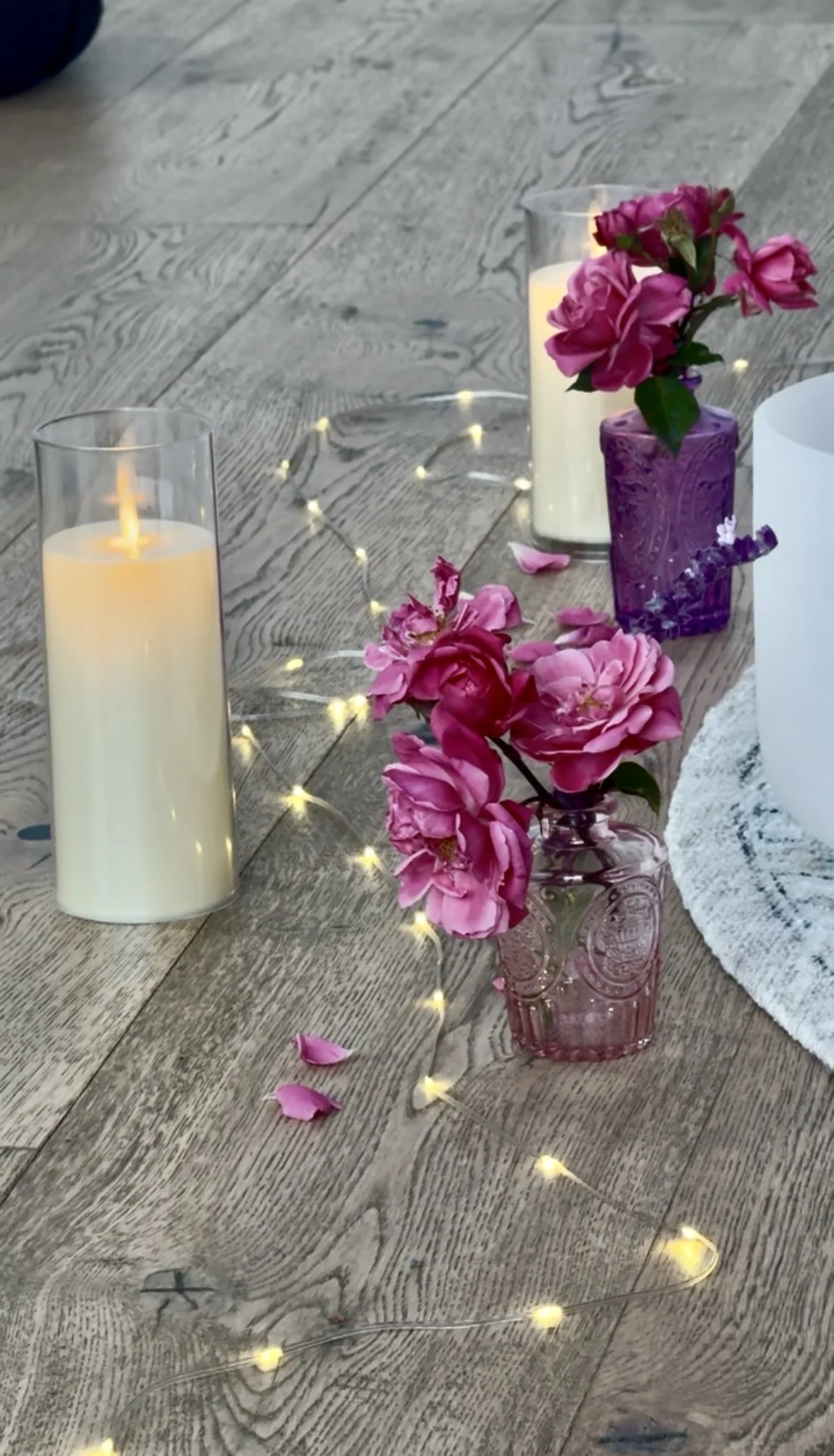 Decorative table setup with pink flowers in glass vases, lit candles in glass holders, a round white centerpiece, and string lights on a wooden surface.