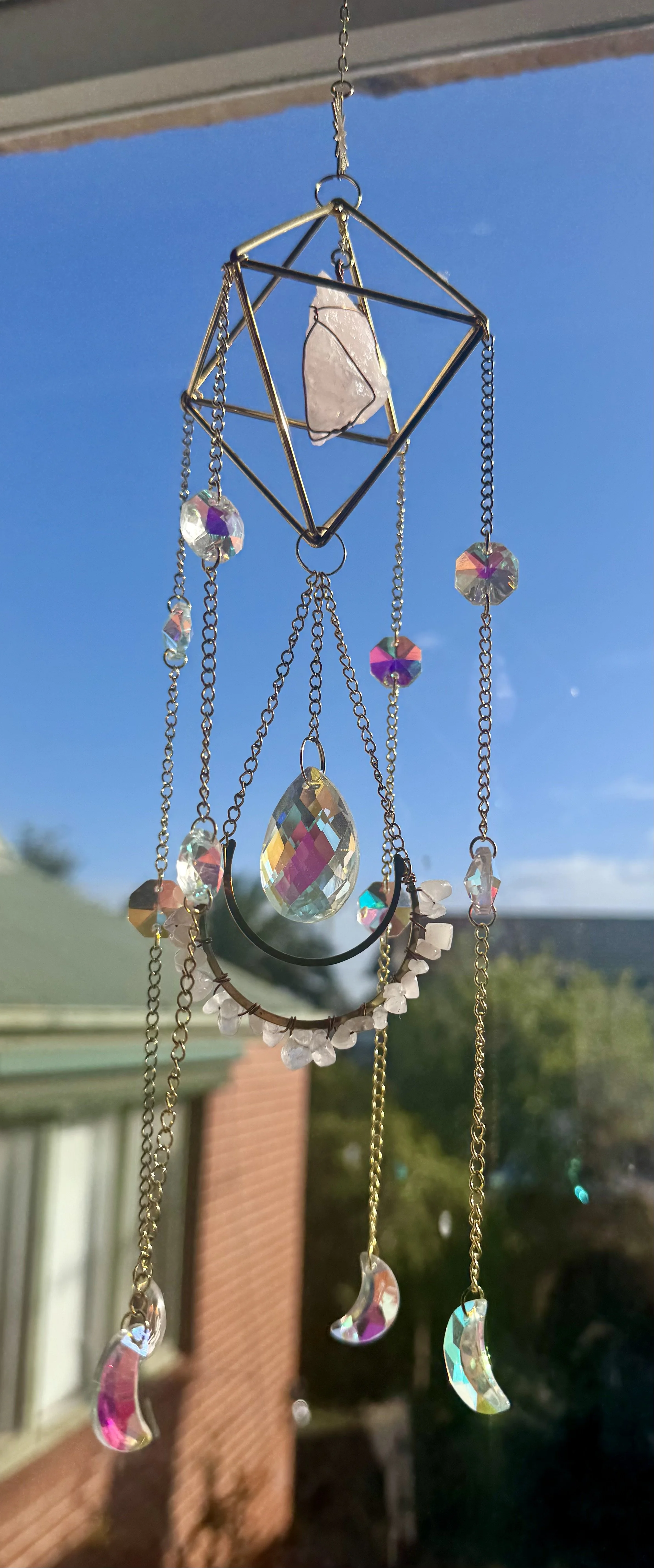 A decorative hanging ornament with chains, crystals, and a geometric metal frame, set against a clear blue sky with part of a house and trees in the background.