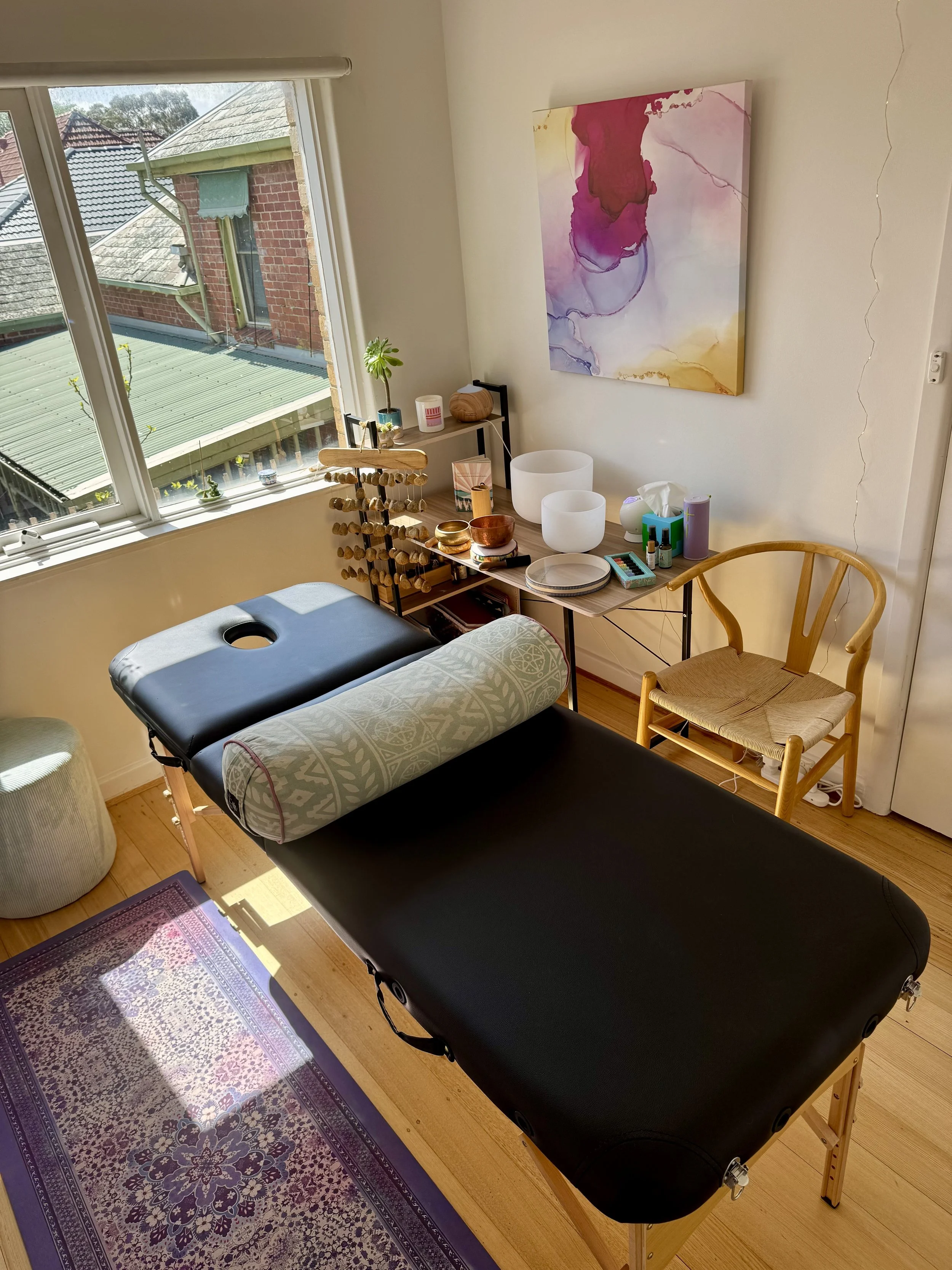 Massage therapy room with massage table, a decorative pillow, wooden chair, and meditative singing bowls. Window showing rooftops outside.