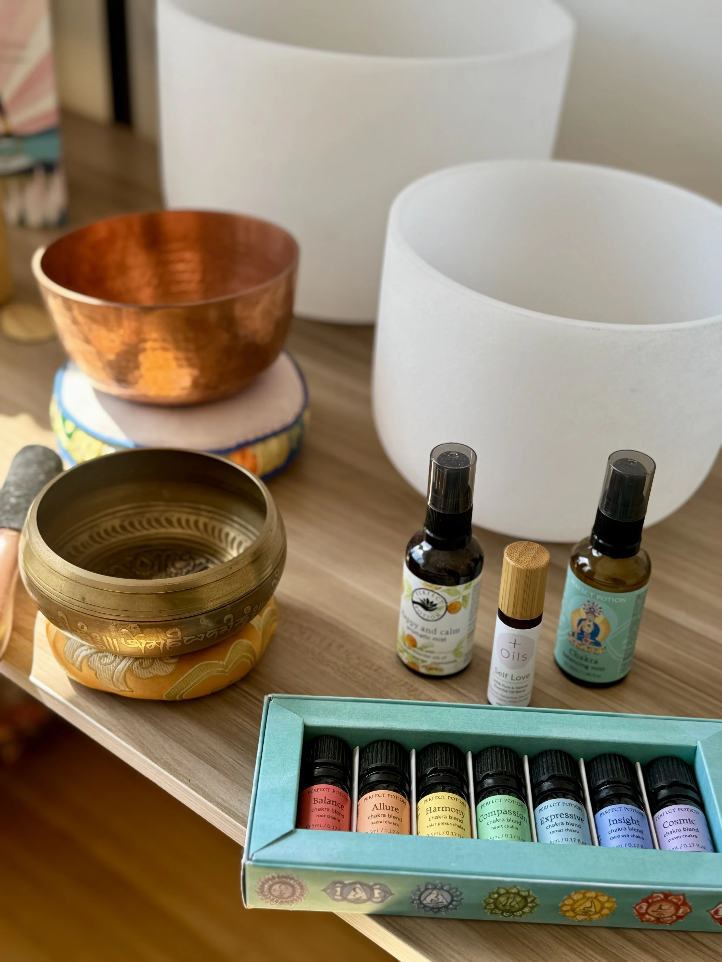 A collection of singing bowls, essential oils, and chakra incense sticks on a wooden table, with white and copper bowls and a colorful box of chakra balancing oils.