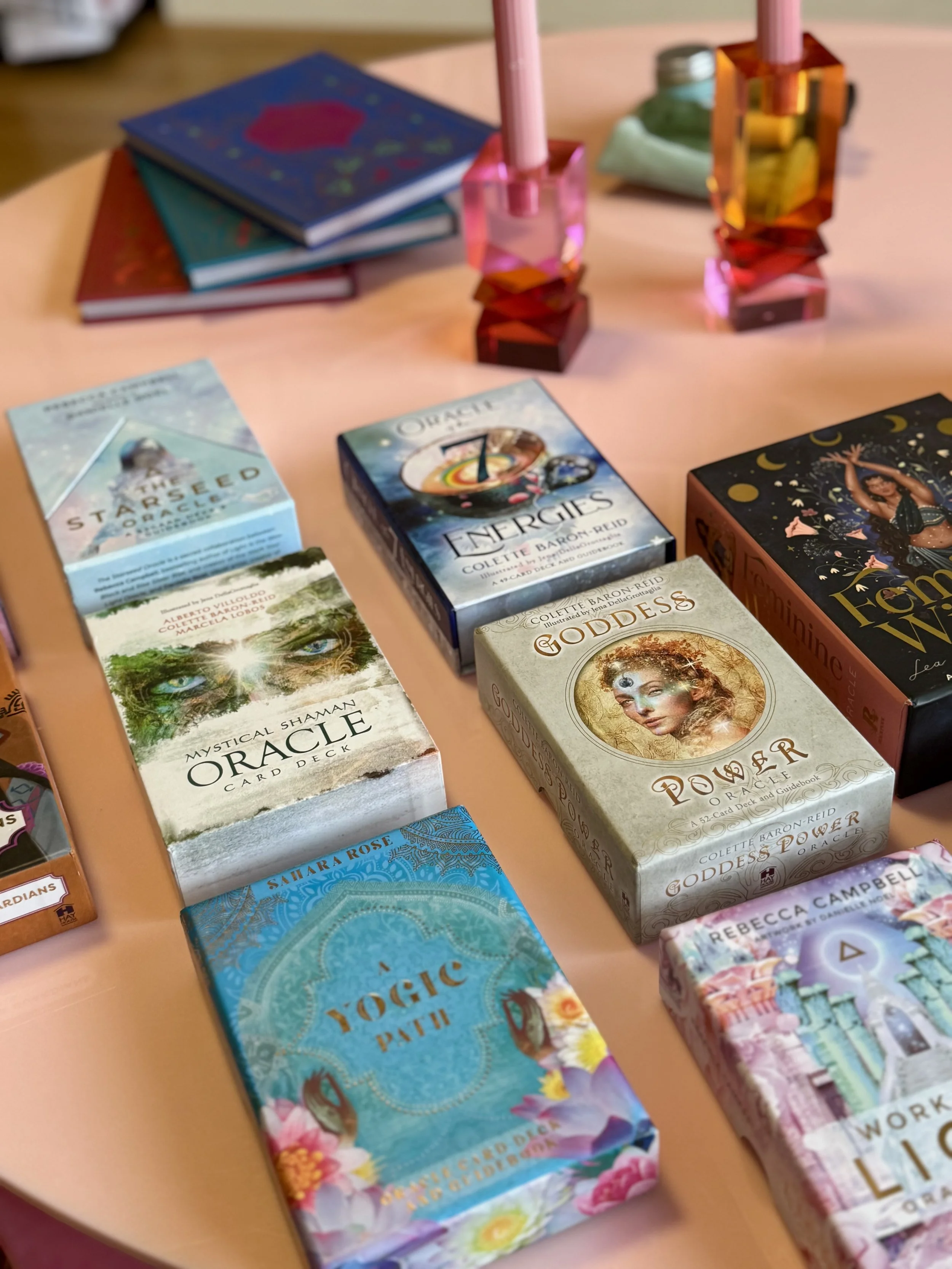 A collection of tarot, oracle, and spiritual cards titled 'Oracle of the Unicorns,' 'Energies,' 'Goddess Power,' 'Lightworker,' 'Mystical Shaman Oracle,' 'Vogue Tarot,' and 'Rebecca Campbell Work Your Light' on a pink table, along with two pink candl