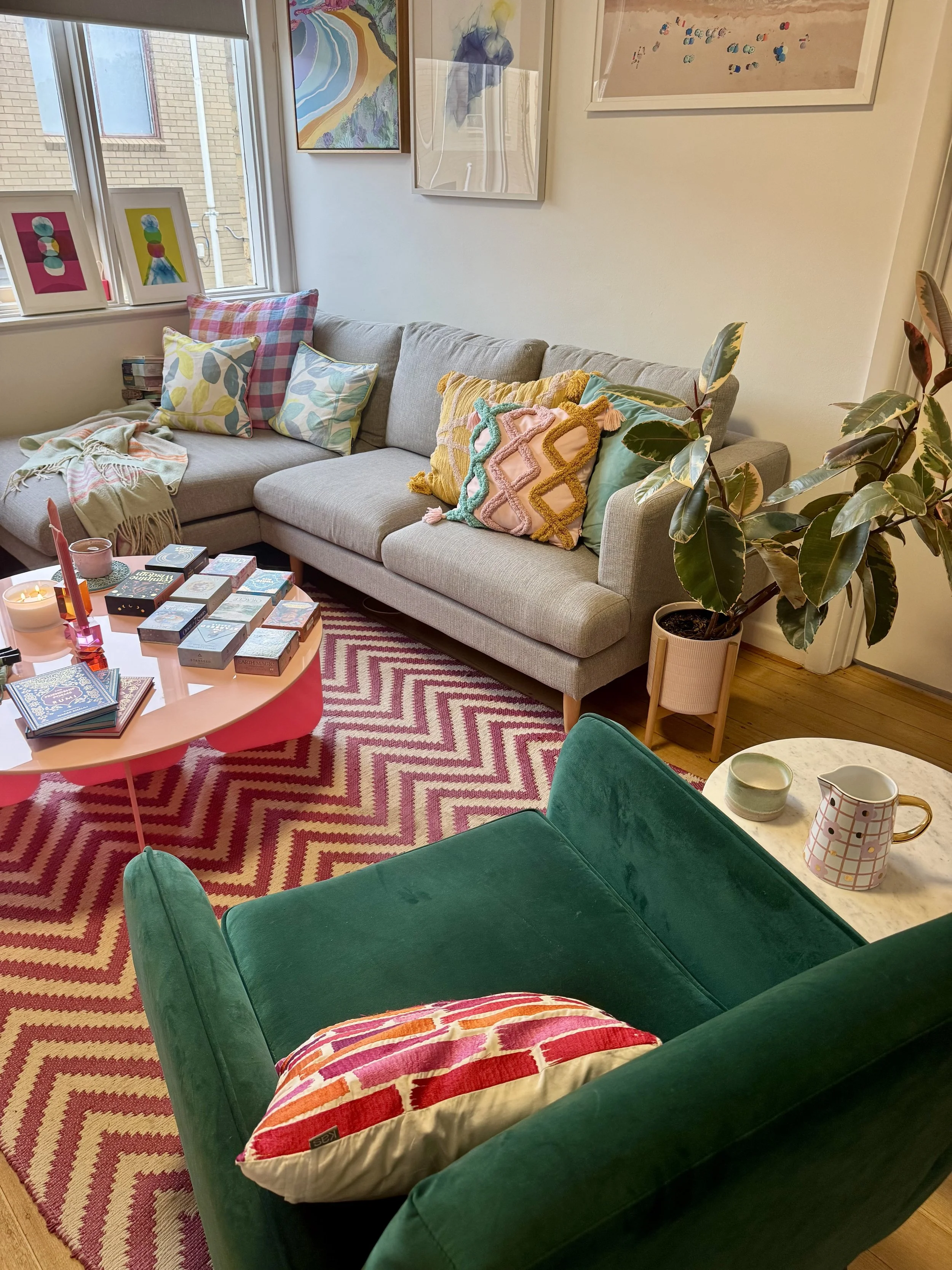 A cozy living room with a gray sofa with colorful pillows and a blanket, a round pink coffee table with books and candles, a green velvet chair, a small marble side table with a pitcher and bowls, a tall plant in a pink pot, and artwork on the walls,