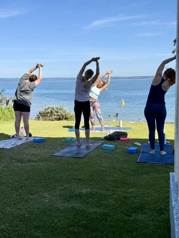 Four women practicing yoga on mats outdoors near a body of water on a sunny day.