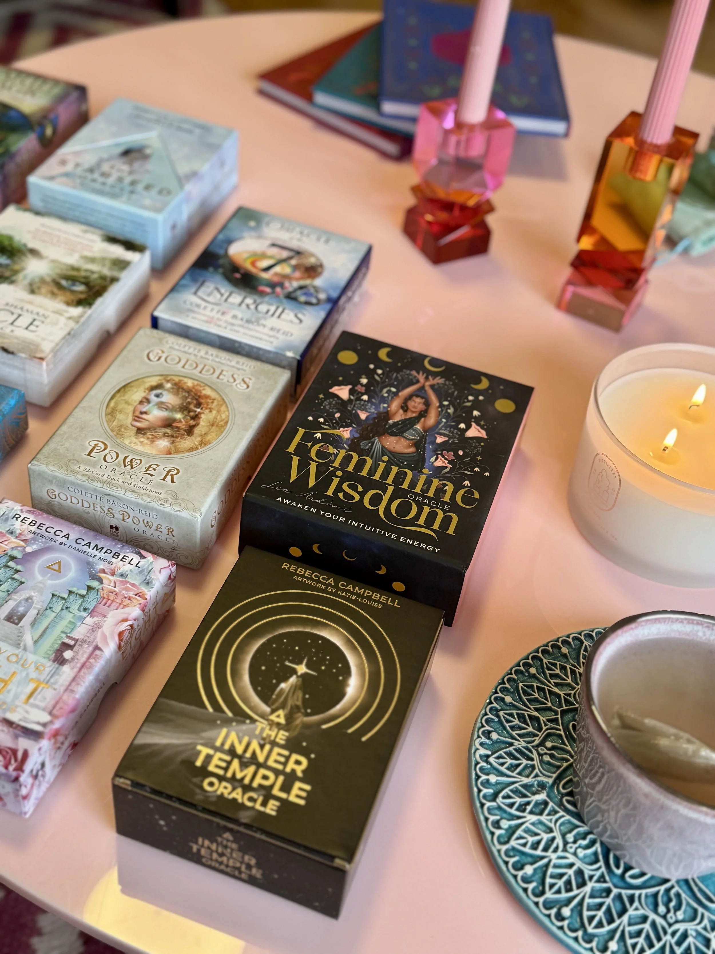Table with various oracle and tarot card decks, including 'Feminine Wisdom', 'Goddess Power', and 'The Inner Temple'. There are pink and orange candles, a lit white candle in a glass container, a decorative plate, and a partially visible cup or bowl