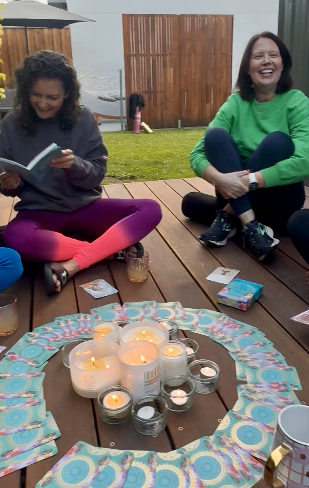 Two women sitting on a wooden deck outdoors, surrounded by candles and tarot cards, enjoying a social gathering with drinks.