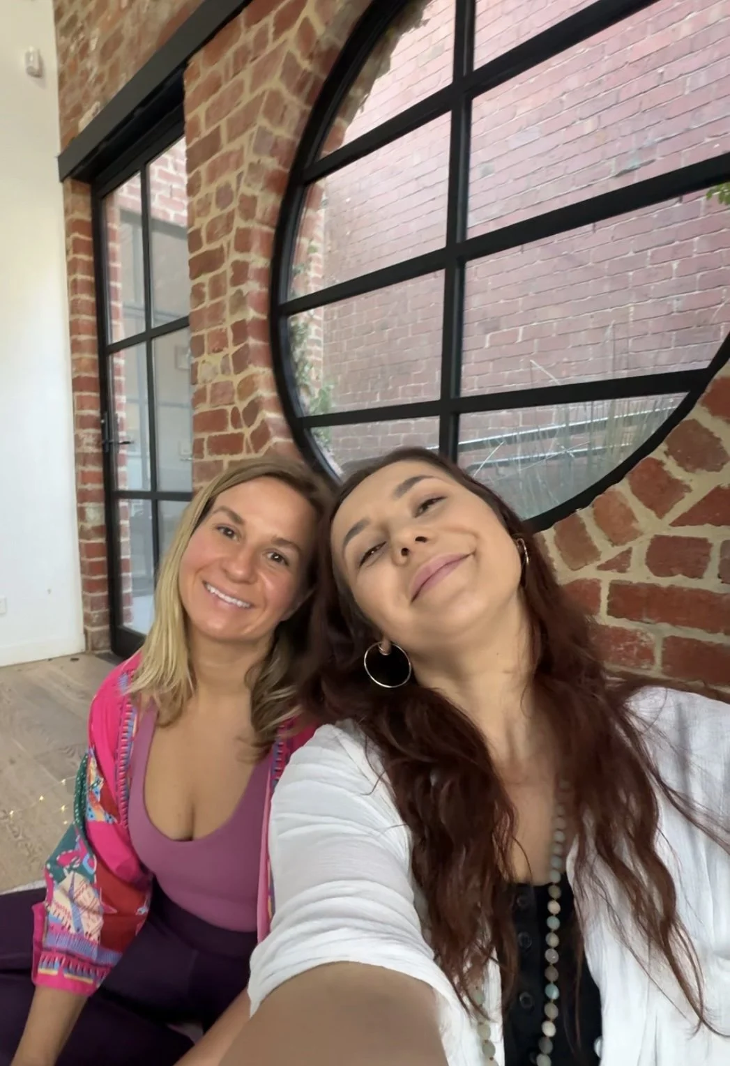Two women taking a selfie together indoors with exposed brick walls and a large window with black framing behind them.