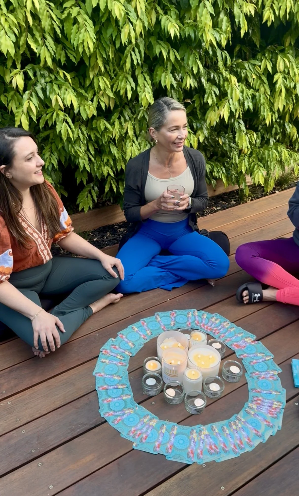 Three women are sitting cross-legged on a wooden deck in front of a large green leafy hedge, participating in a meditation or spiritual gathering, with a circle of lit candles and tarot cards on the ground in front of them.