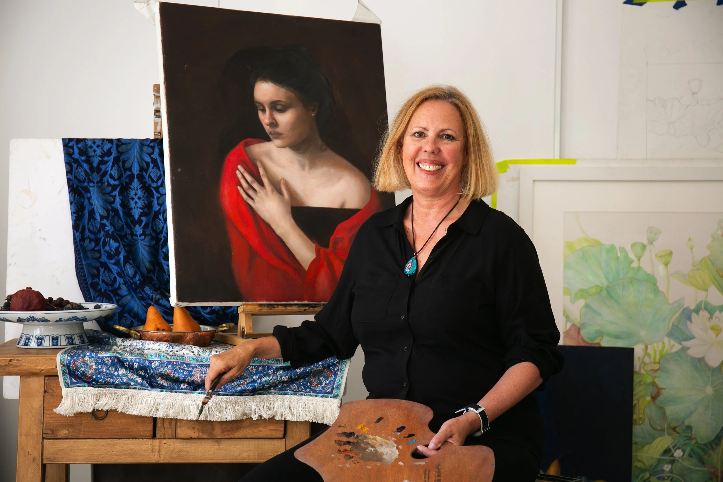 Bronwyn Van de Graff sits in her studio with an easy, welcoming smile, captured mid practice with a brush and palette in hand. Dressed in a simple black shirt she becomes a calm focal point of a space that feel lived in and creative.