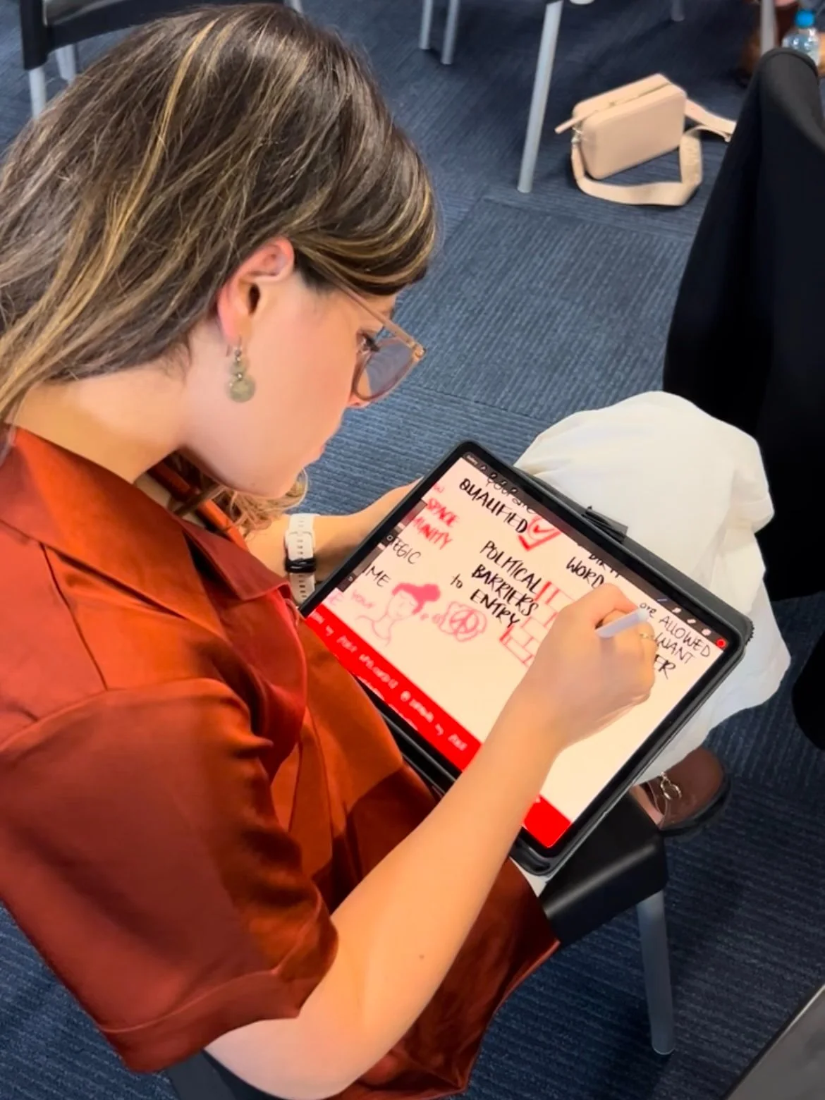 Person in red shirt illustrating infographics on a tablet while sitting in a room with a blue carpet.