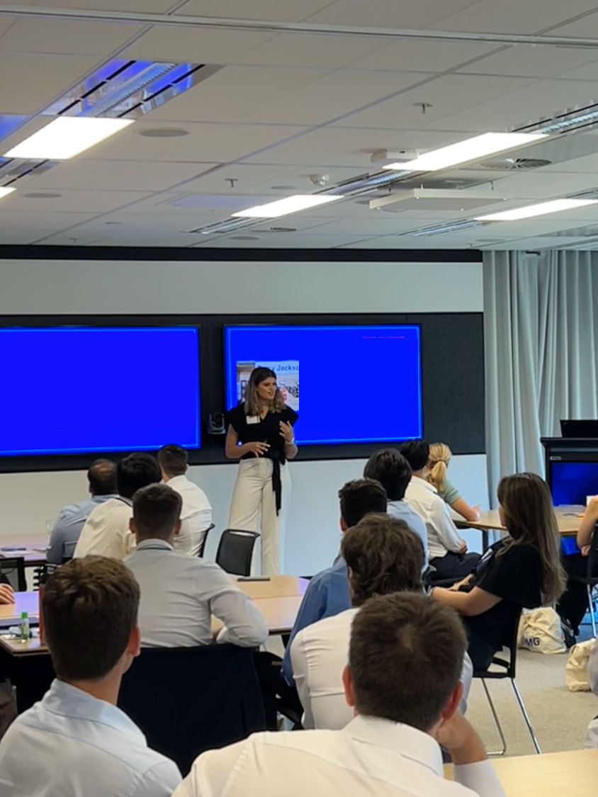 A woman is giving a presentation in a classroom setting, with several people seated at tables. There are two large screens behind the presenter displaying blue screens. The room is modern with bright lighting and a ceiling with fixtures.