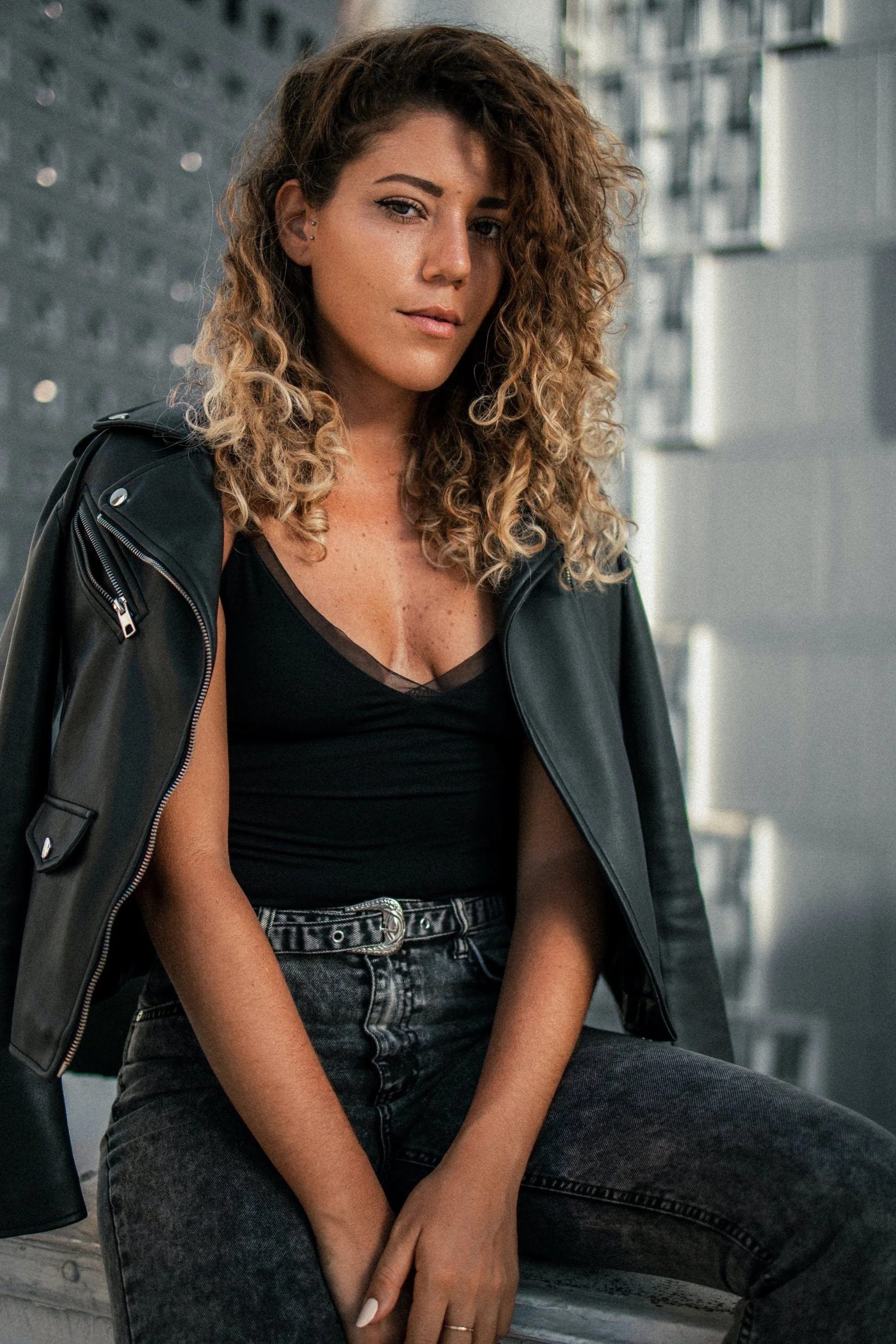 Young woman with curly hair wearing a black top and leather jacket, sitting outdoors
