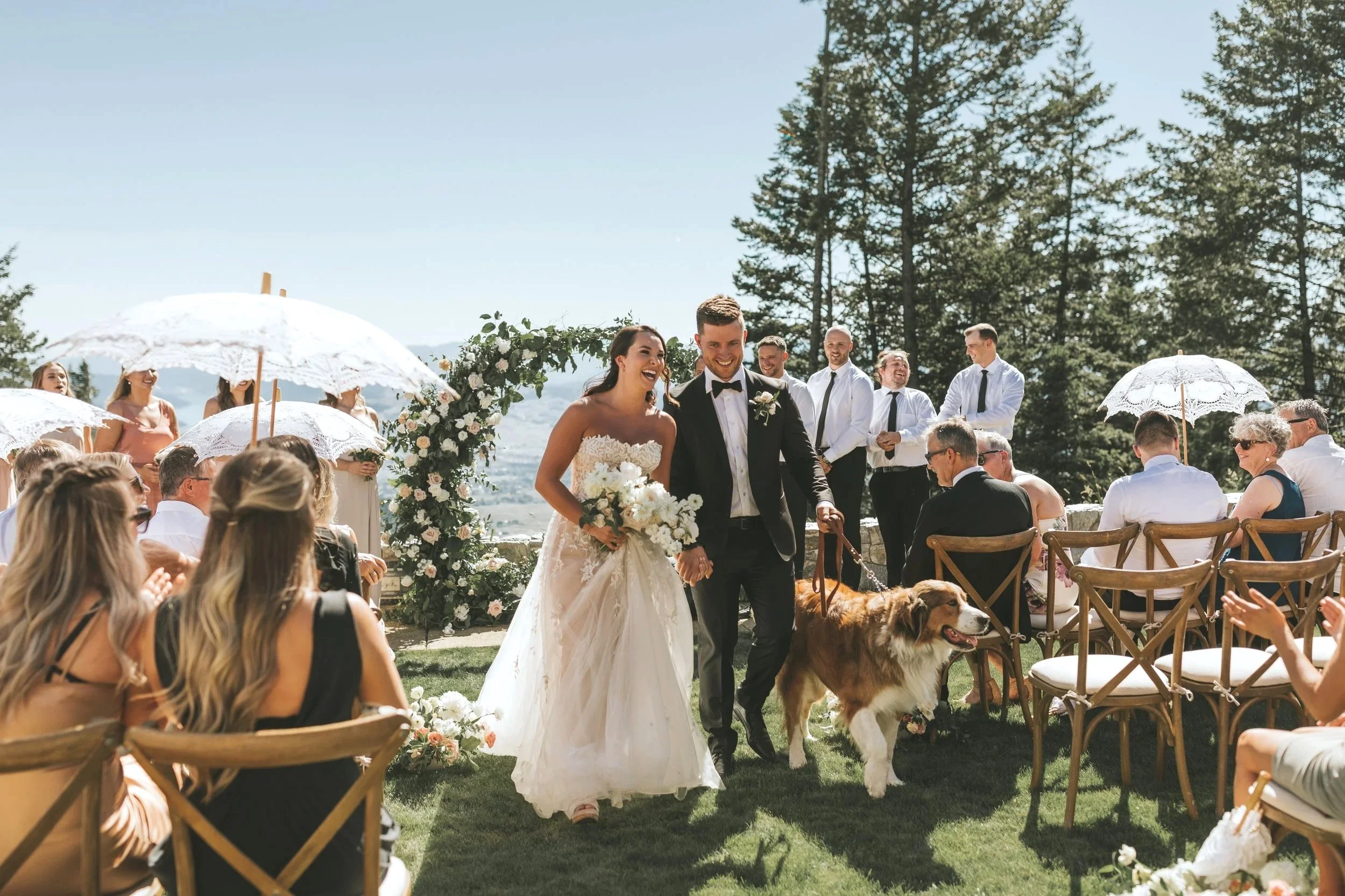 A bride and groom share an intimate moment in a natural outdoor setting, with the groom kissing the bride on her forehead, both smiling softly.