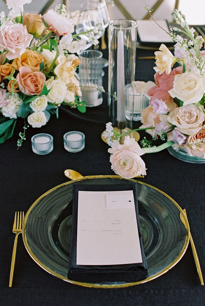 Elegant table setting with black tablecloth, gold-rimmed glass plate, gold utensils, floral centerpiece with pink, white, and peach roses, glass candle holders, and tall candles.