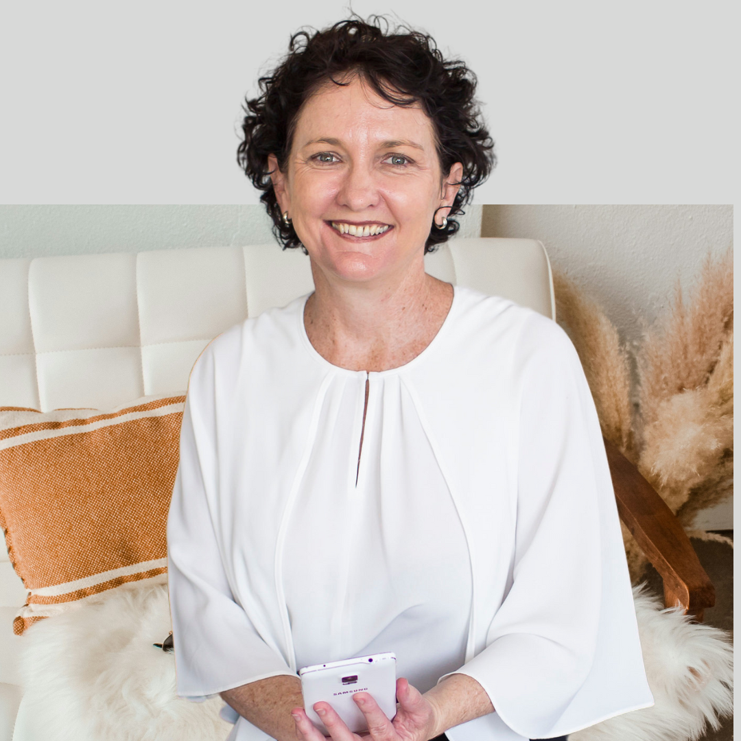 A smiling woman with short curly dark hair, wearing a white blouse, sitting on a white chair with a beige and orange pillow and beige pampas grass in the background, holding a smartphone.