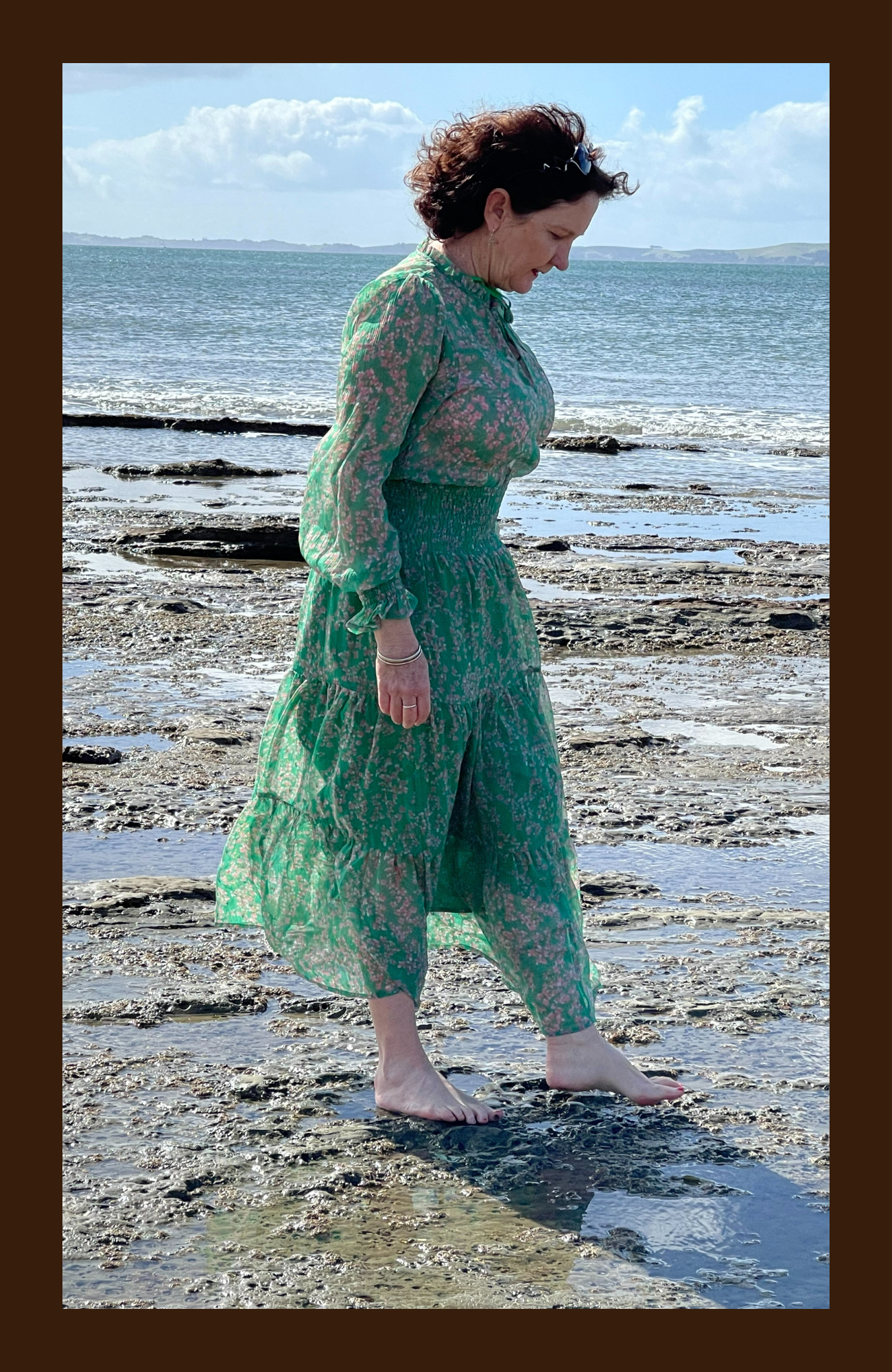A woman in a green floral dress walking barefoot on a rocky beach near the ocean during the daytime.