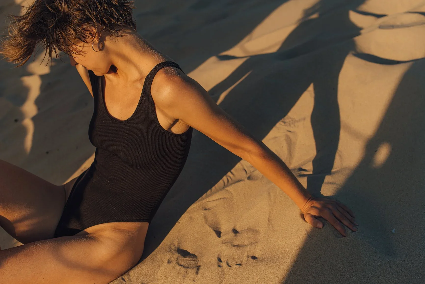 A woman in a black swimsuit sitting on the sand at a beach during sunset, casting a long shadow.