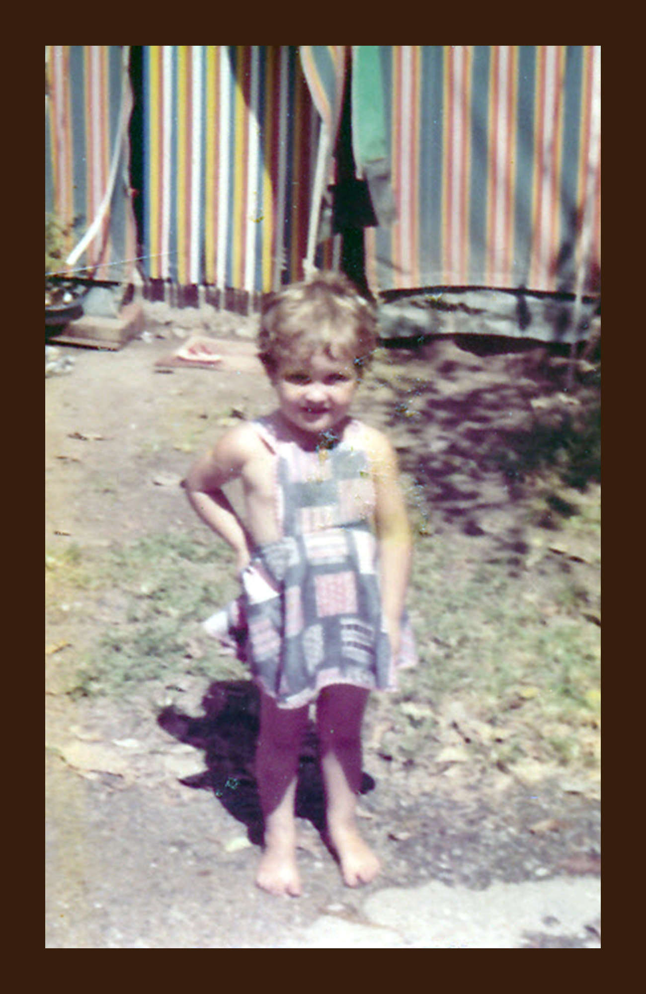 A young girl standing outdoors on a dirt surface, wearing a colorful dress and maroon tights, with a striped fabric background in the distance.