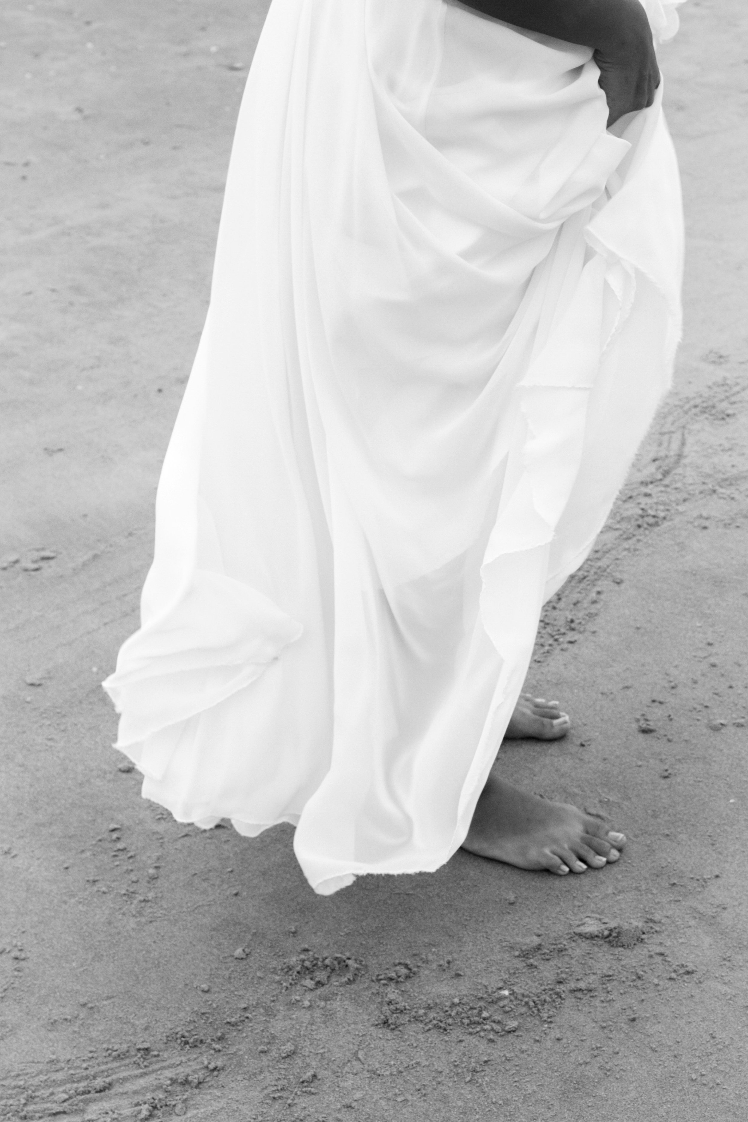 A person standing barefoot on sandy ground, wearing a flowing white dress or robe.
