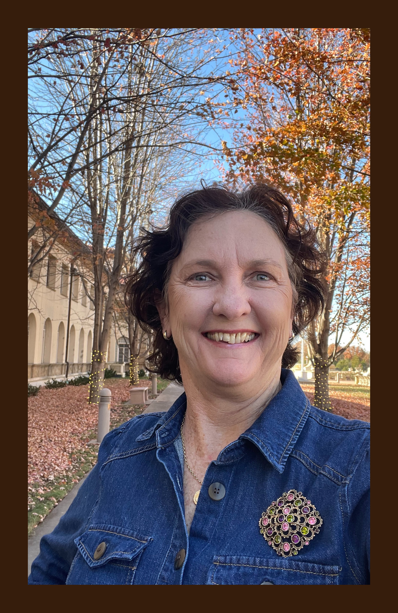 Smiling woman taking a selfie outdoors in autumn, wearing a denim jacket with a large brooch, with trees with fall foliage and a historic building in the background.