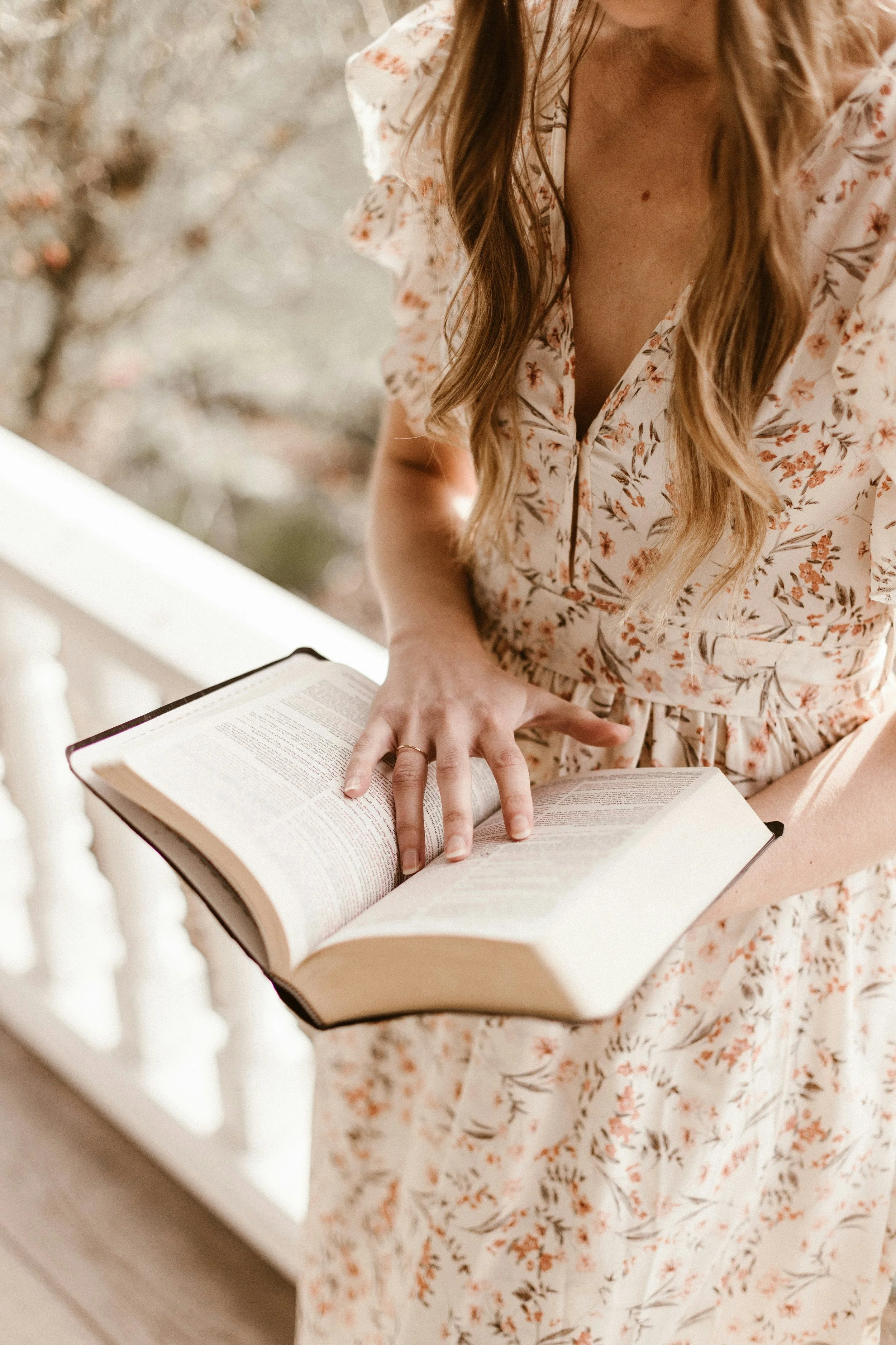 A woman wearing a floral dress is reading a large open Bible on a porch or balcony with a blurred background of trees and blossoms.