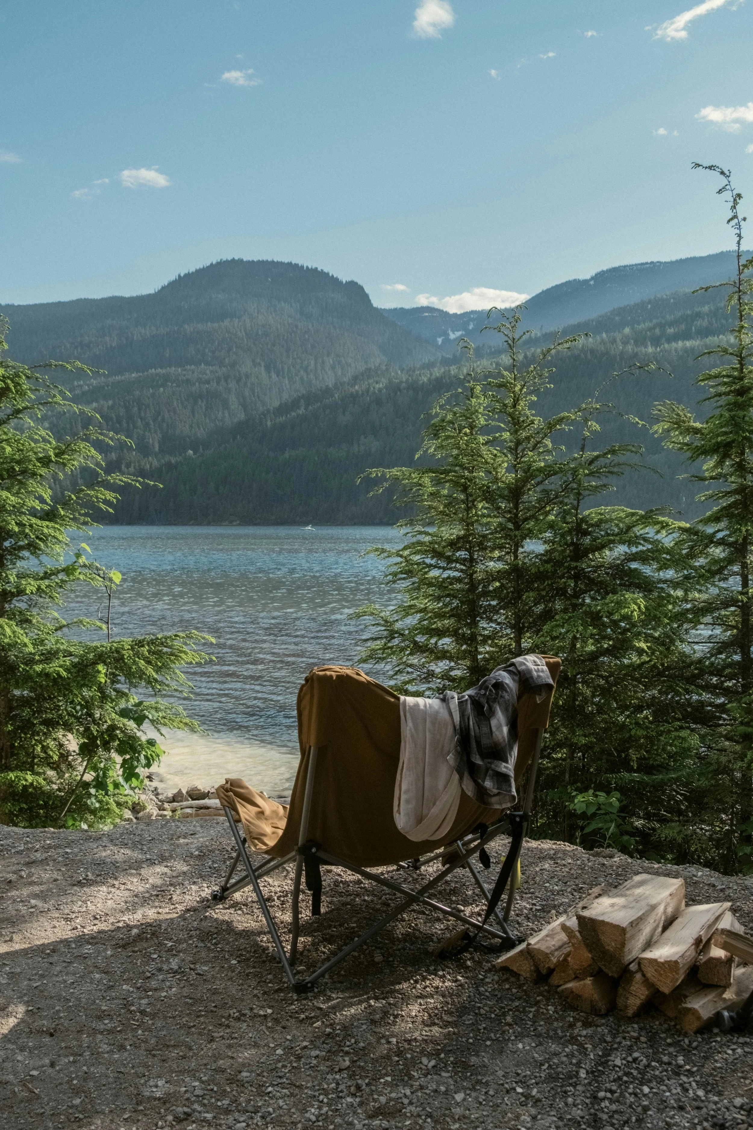 A camping chair on a dirt ground by a lake with surrounding pine trees and mountains under a partly cloudy sky.