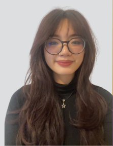 Young woman with long wavy hair, glasses, and a star necklace smiling, dressed in a black top against a light background.