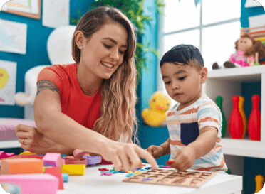 A woman and a young boy playing with colorful clay and toys at a table in a bright, cheerful room.