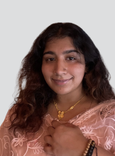 Portrait of a young woman with dark, curly hair wearing a pink saree and gold jewelry, smiling slightly.