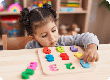 A young girl with dark hair in two buns is sitting at a table playing with colorful foam number puzzles.