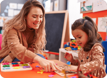 Woman and young girl playing with colorful puzzle pieces at a table in a classroom or playroom.