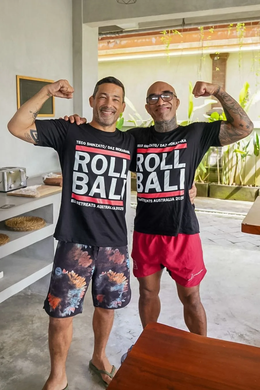 Two men flexing their biceps while smiling, wearing matching 'ROLL BALI' t-shirts, standing in a room with plants and a patio outdoor area.