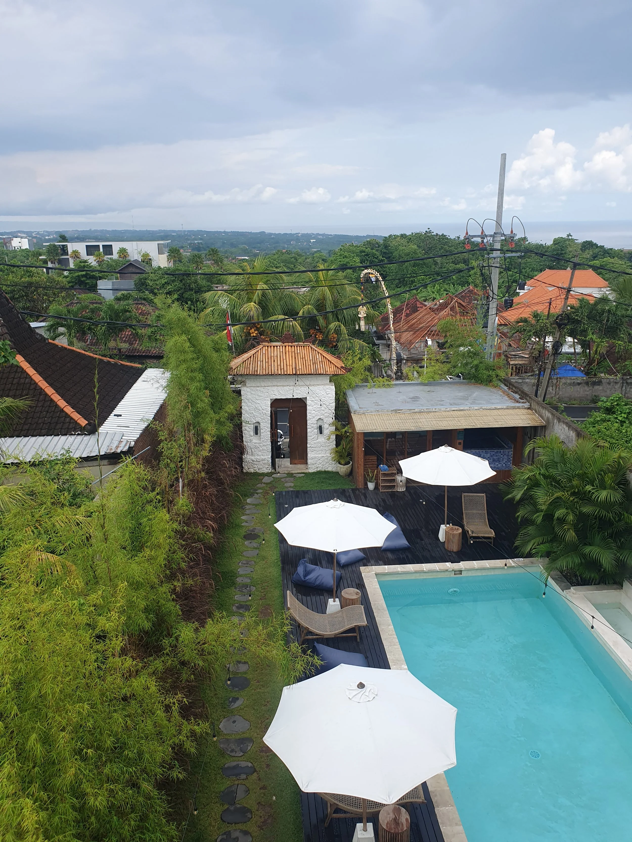A backyard with a swimming pool, white umbrellas, lounge chairs, lush trees, and a view of rooftops and distant landscape under cloudy skies.