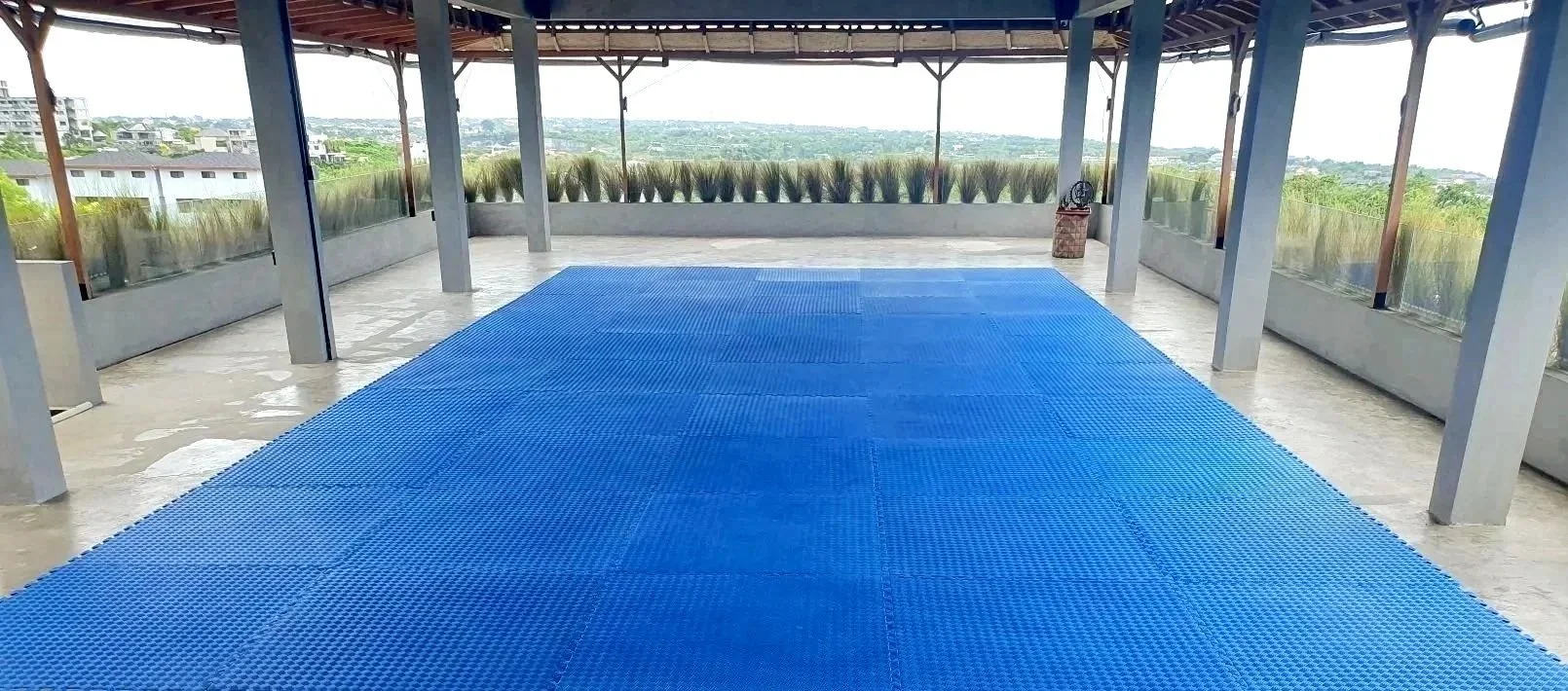 Rooftop terrace with a blue rubberized sports surface, surrounded by glass railings, with a scenic view of nearby buildings and greenery.
