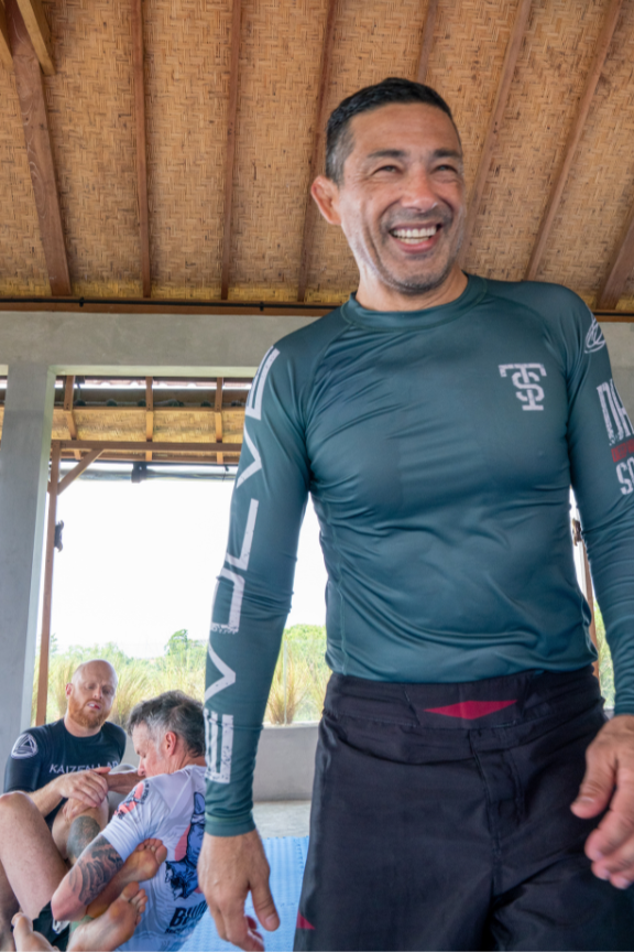 A BJJ coach smiling and wearing a teal long-sleeve no-gi rash guard while coaching a class.